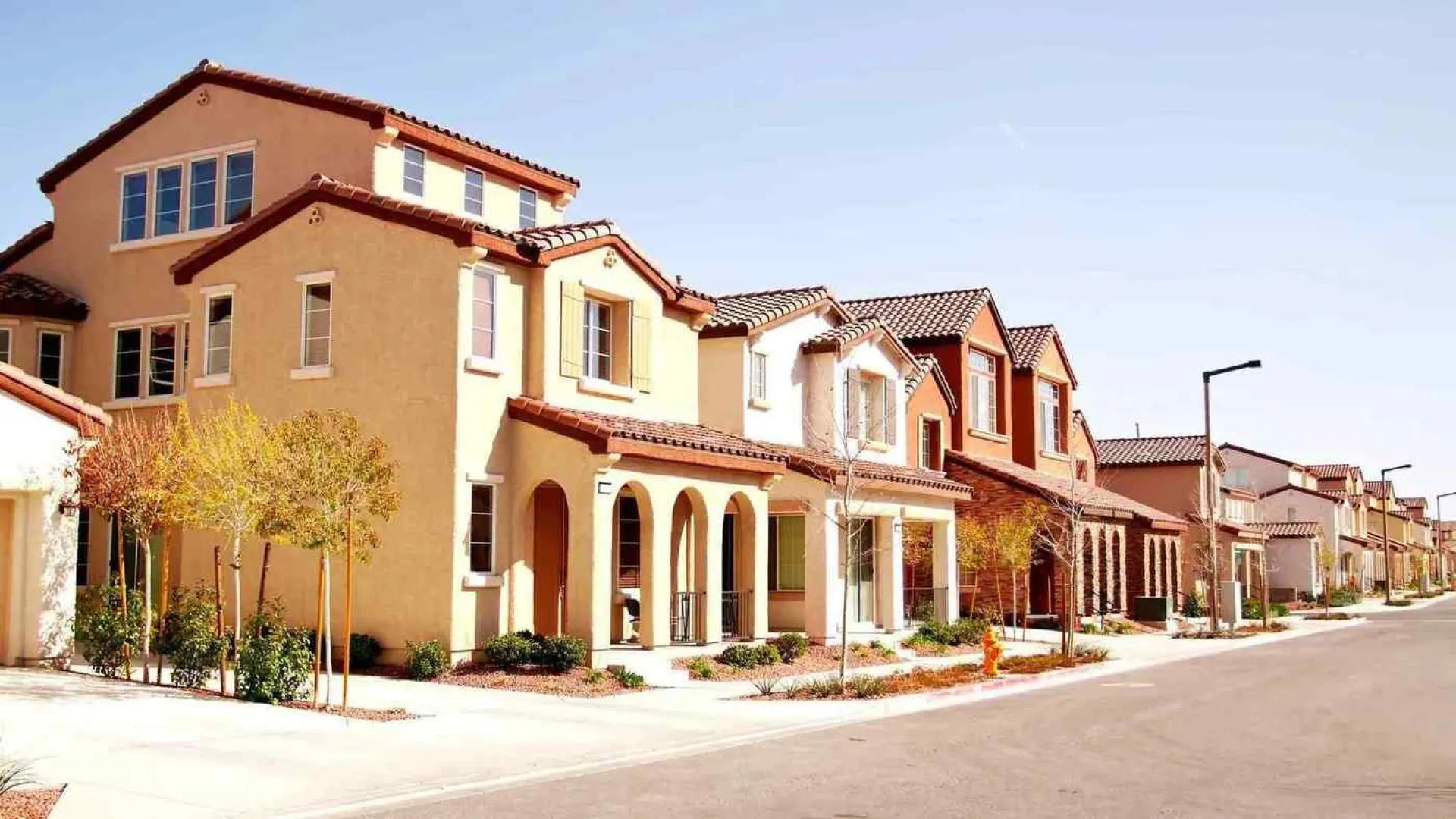 Sunny suburban street with modern Mediterranean-style homes, trees, and a clear blue sky.