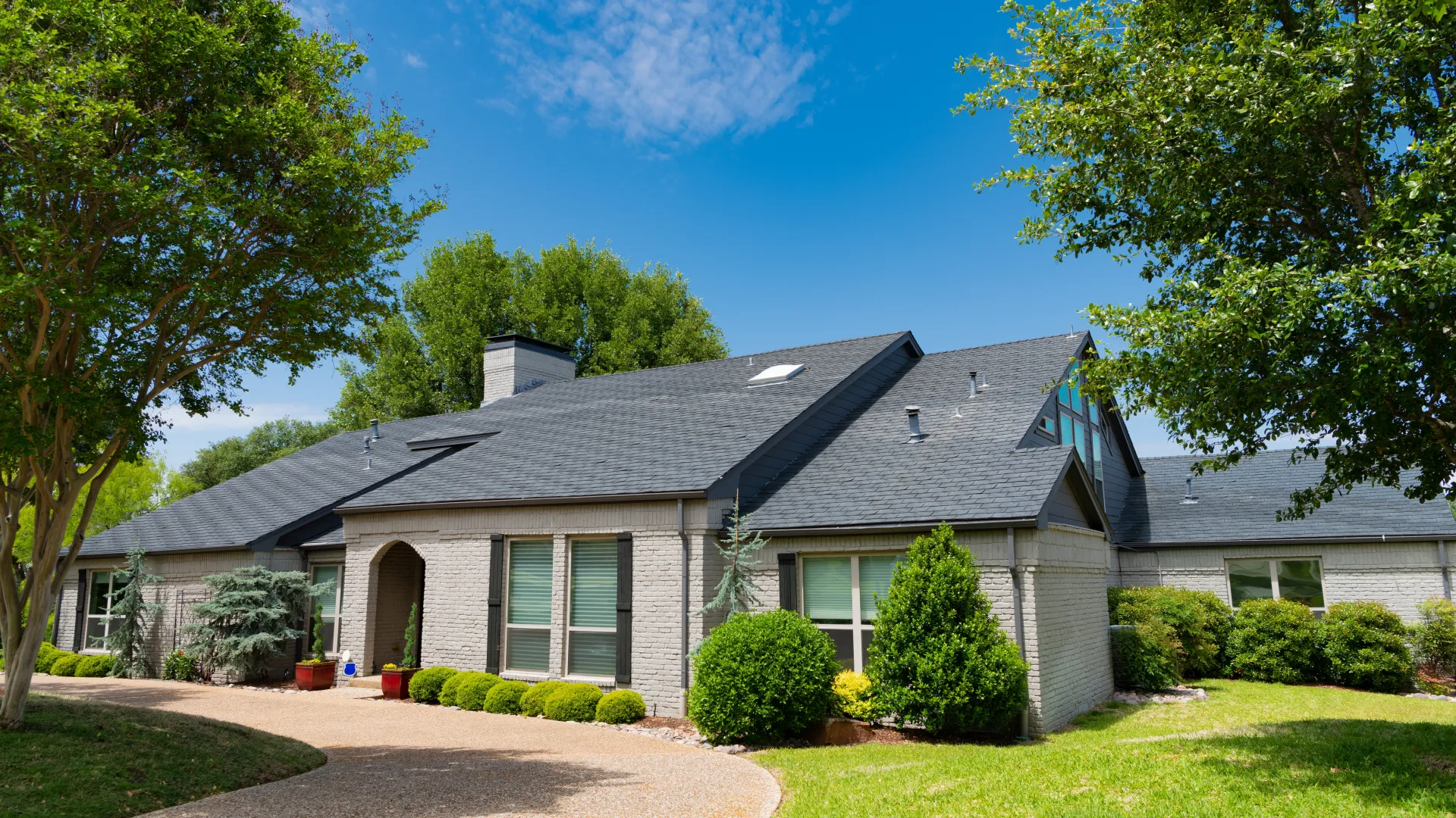 Modern single-story house with gray brick exterior, dark roof, and surrounding green trees and lawn under blue sky.