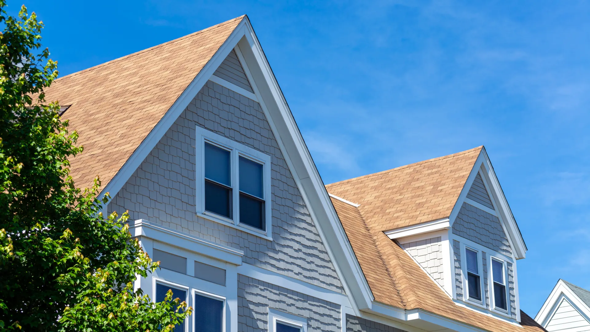 Gray house with steep gable roof and tan shingles against a clear blue sky on a sunny day