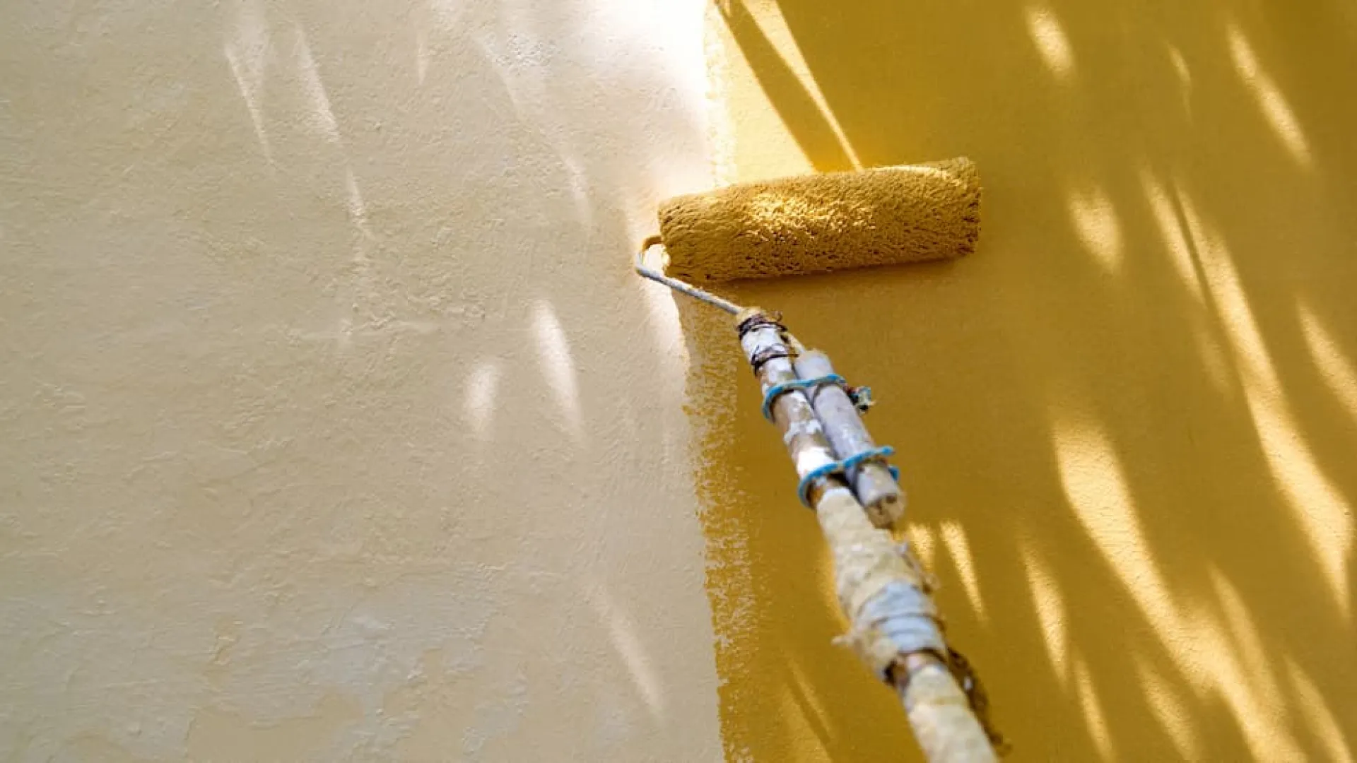 Paint roller applying fresh yellow paint on a wall in bright natural light with shadows