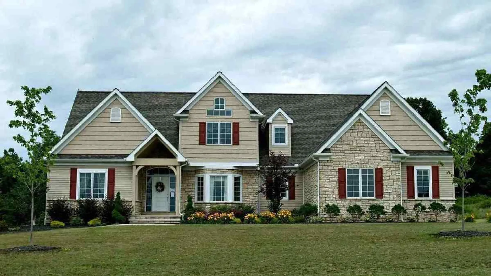 Spacious two-story suburban house with beige siding, stone facade, red shutters, and landscaped front yard.
