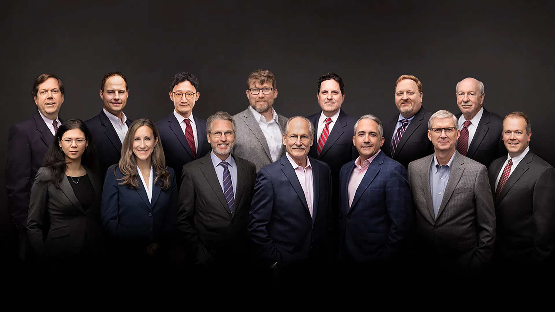 Group portrait of fourteen professionally dressed business executives against a dark background.