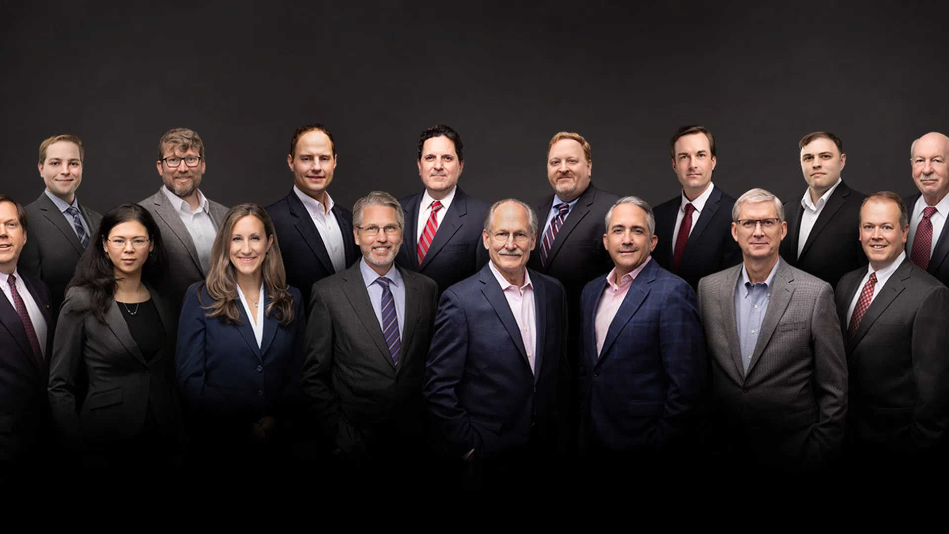Group portrait of 15 business professionals dressed in formal suits against a dark background.