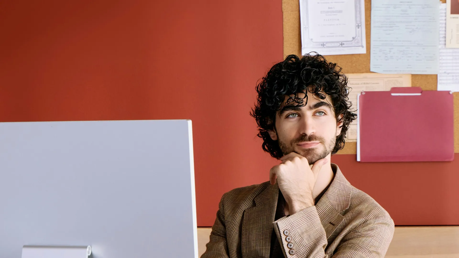 Young man in plaid suit thoughtfully working at a computer desk with red wall and organized corkboard behind
