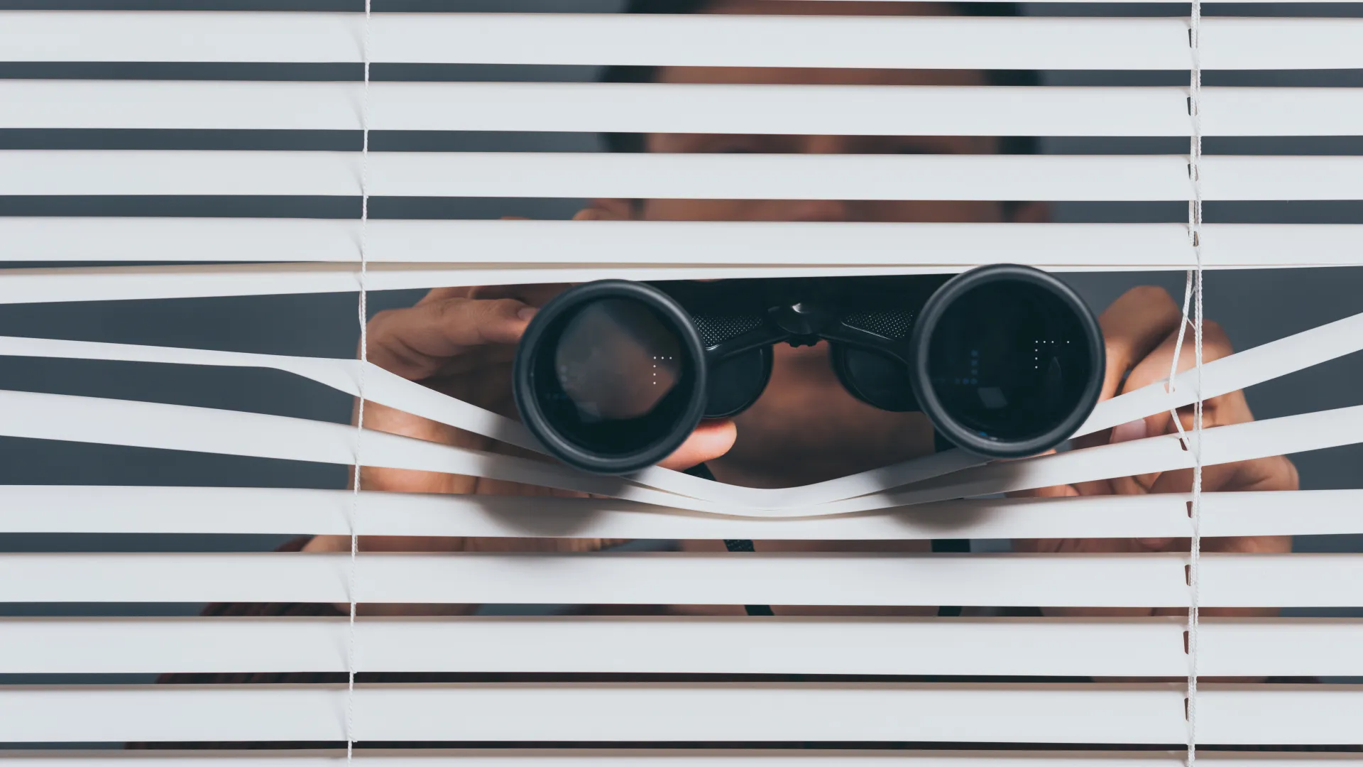 Person looking through white window blinds using black binoculars in a dimly lit setting