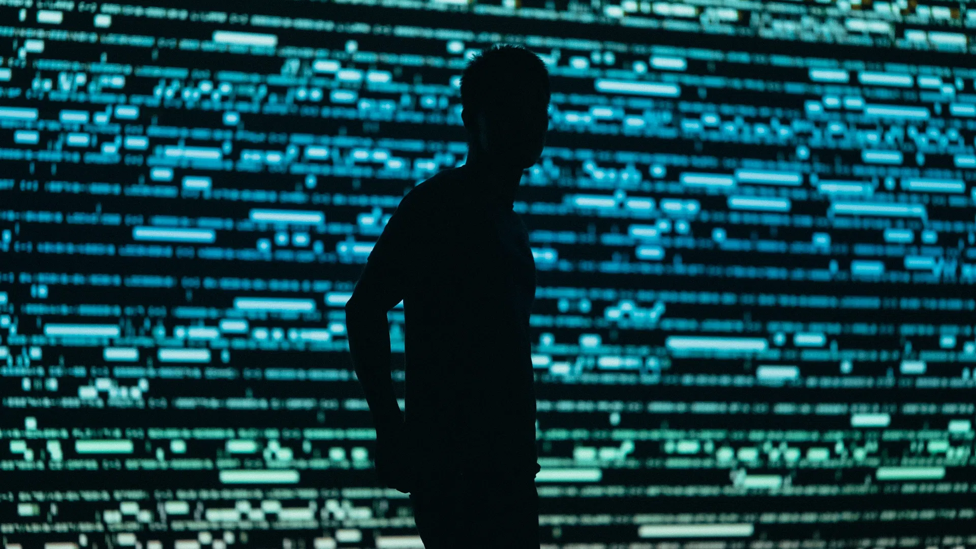 Silhouette of a person standing against a large digital screen filled with blue code and data lines.