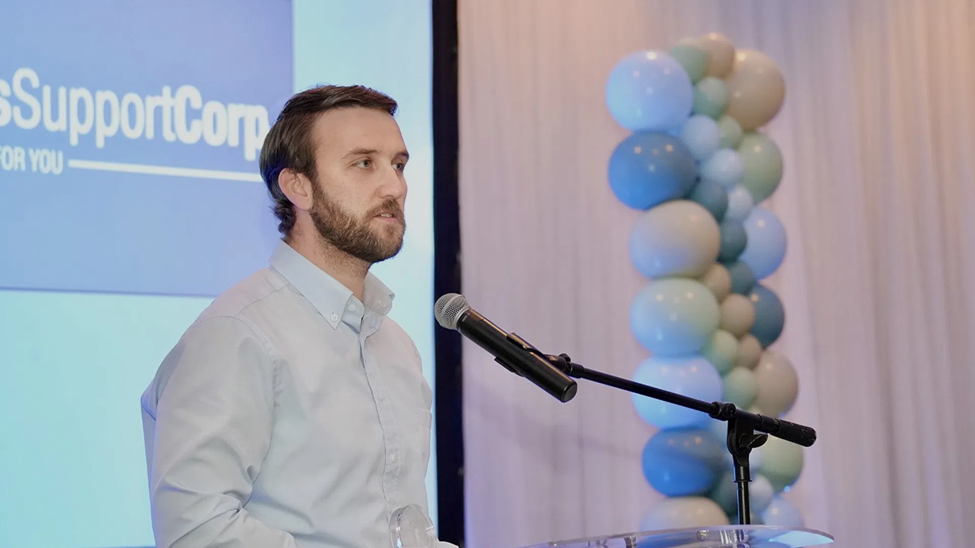 Man in light blue shirt speaking at clear podium with microphone and balloon decoration in background. Photo Courtesy Cape & Plymouth Marketing