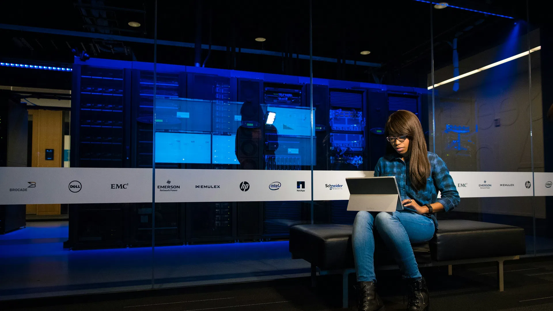 Woman with long hair using a laptop while sitting on a bench in a server room with blue lighting.