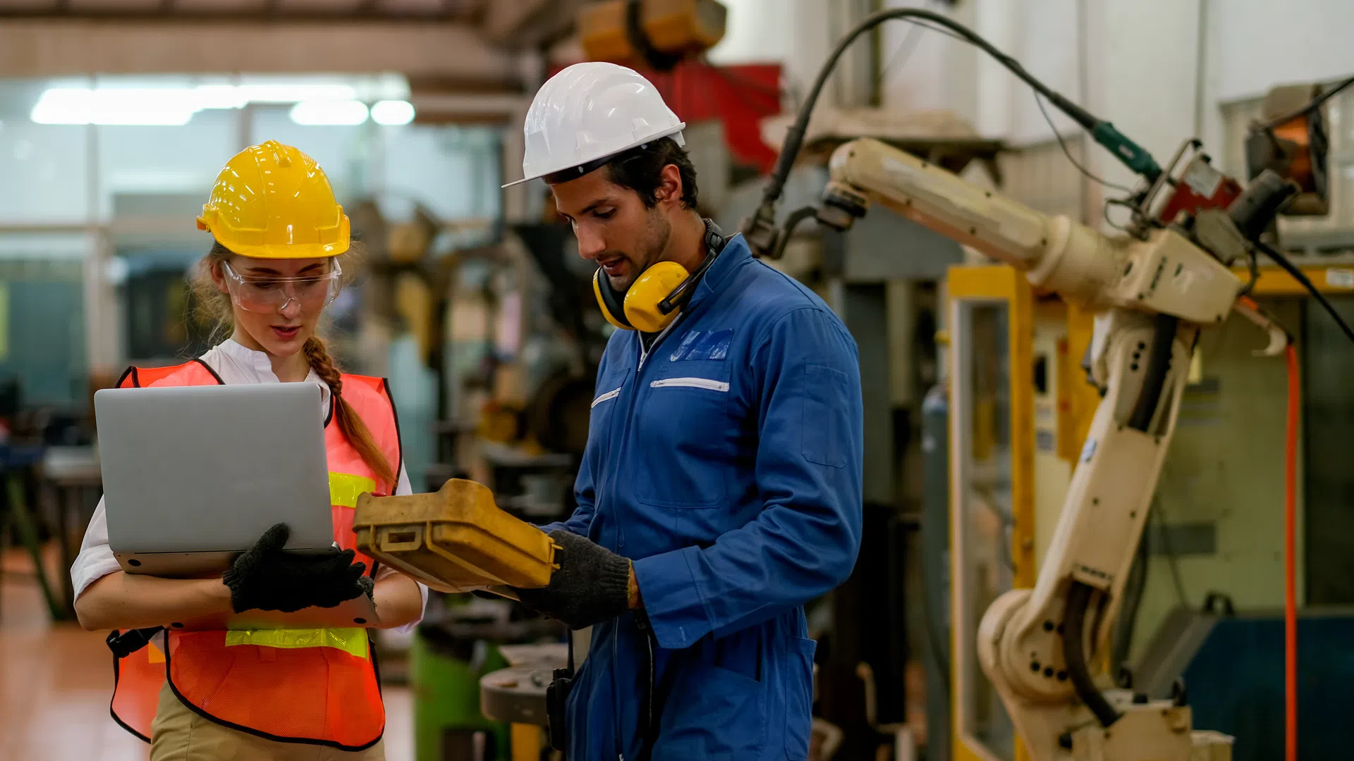 Two engineers wearing safety gear review data on a laptop and equipment in a factory with robotic machinery.