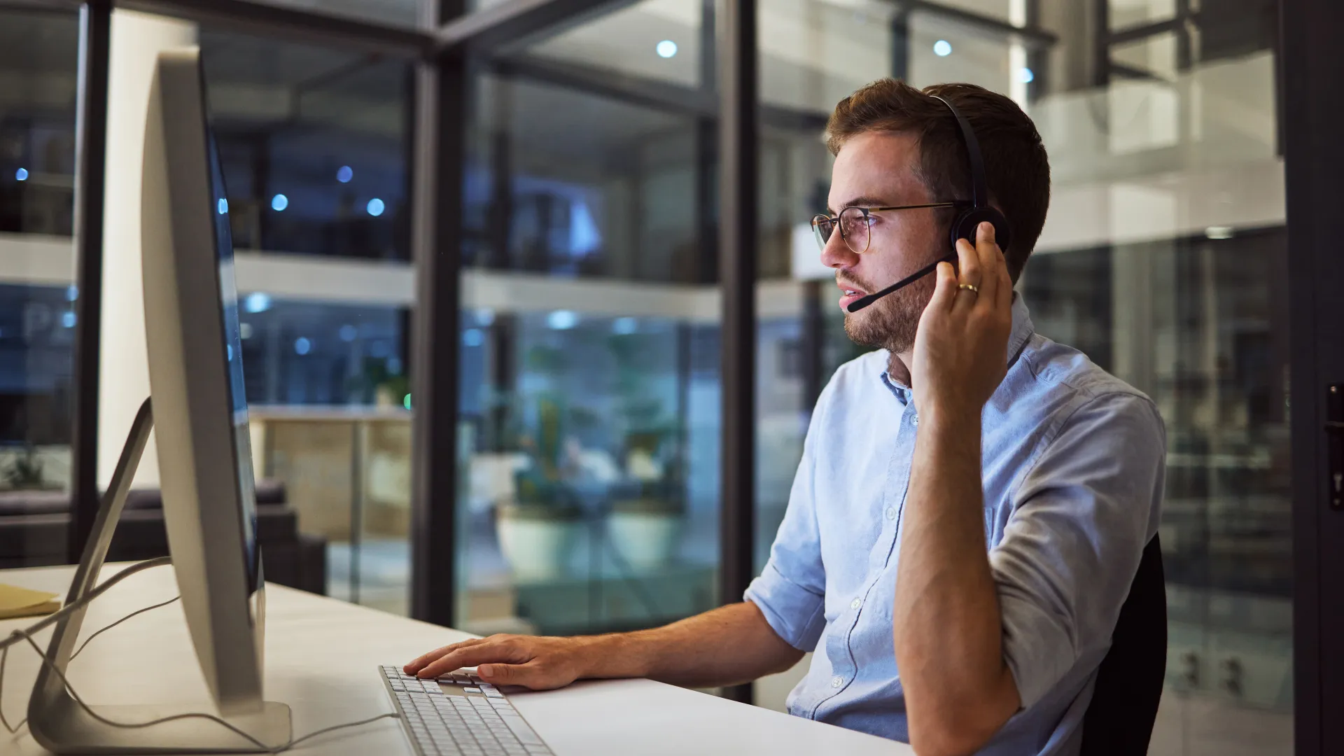 Man wearing headset and glasses working at desktop computer in modern office during evening.