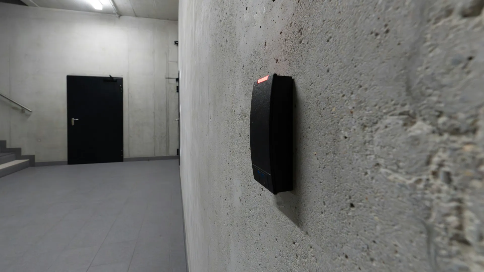 Security card reader mounted on a concrete wall in a minimalistic, industrial stairwell with a black door.