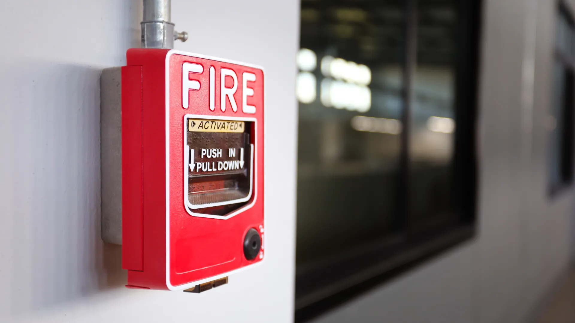 Red fire alarm pull station mounted on a white wall inside a building with a window in the background.