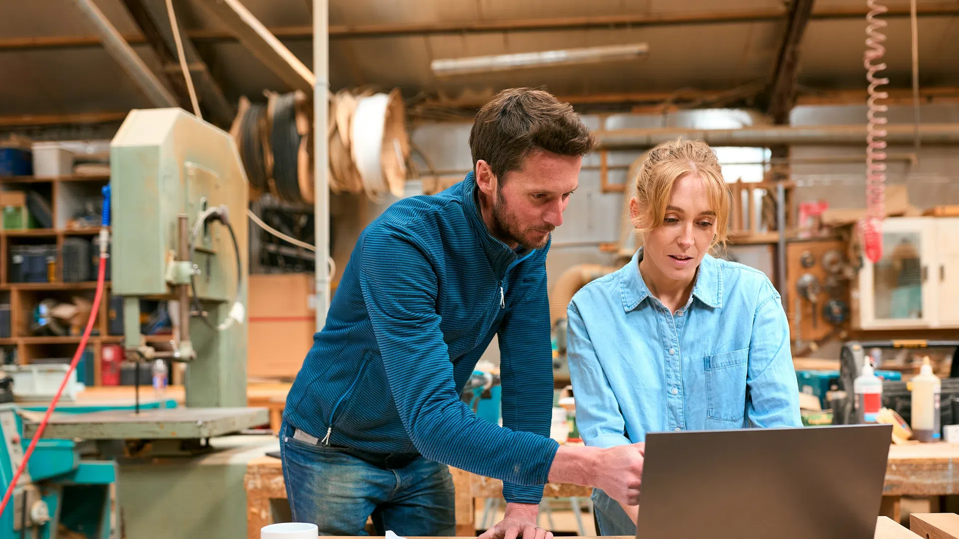 Man and woman collaborating on laptop in woodworking shop surrounded by tools and materials.