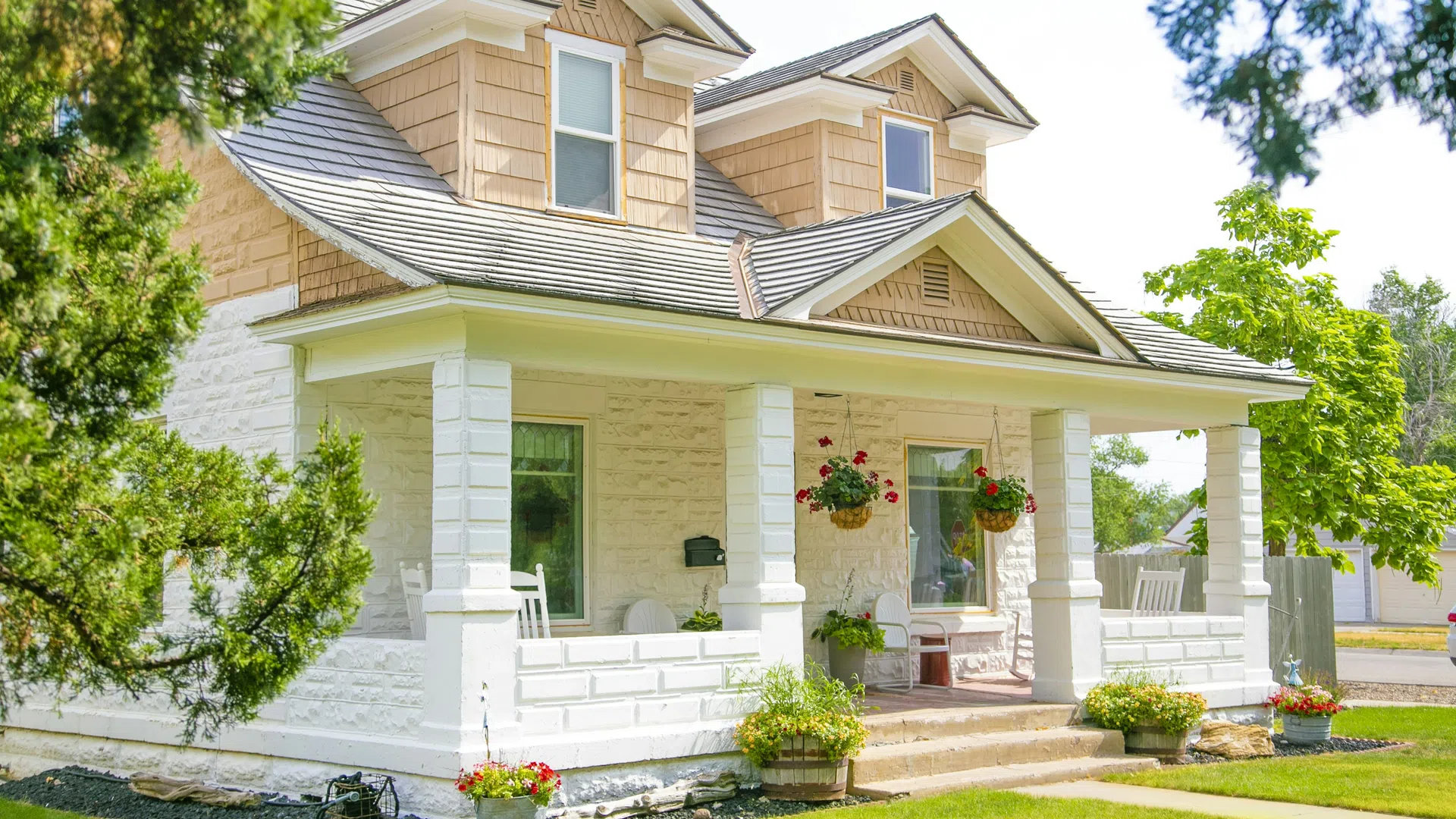 Sunny front view of a charming white stone house with tan shingles, porch swing, and vibrant hanging flower baskets