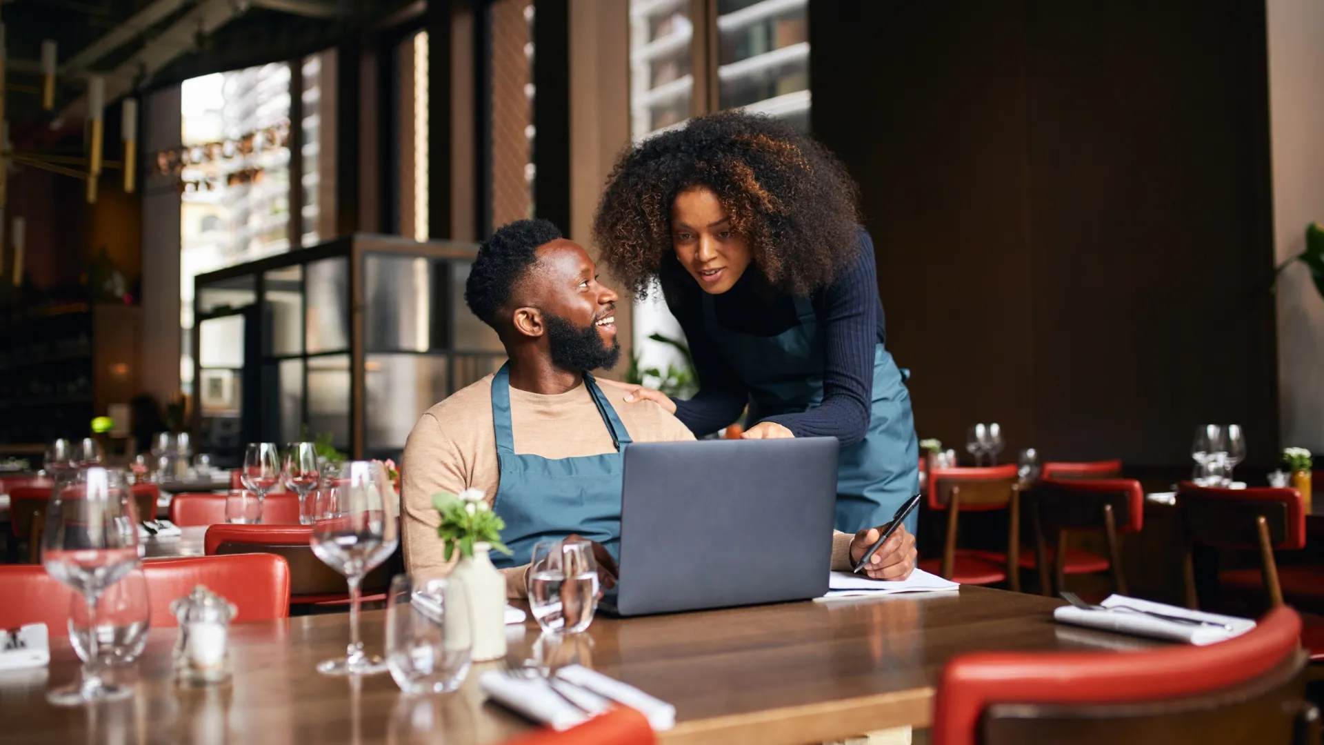 Two restaurant staff in aprons smiling and working together on a laptop in a dining area with wooden tables and red chairs.