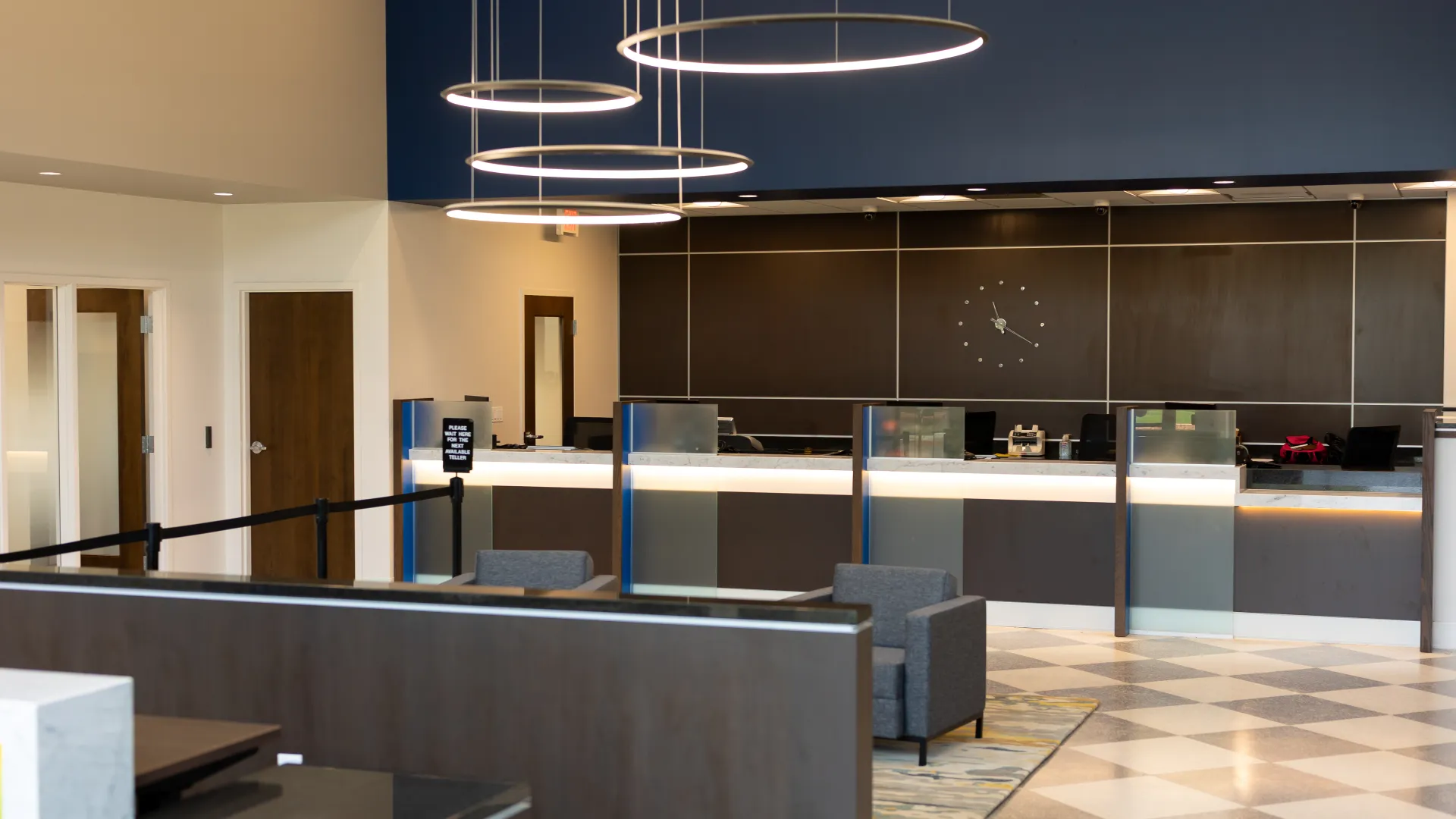 Modern bank lobby with reception desks, circular hanging lights, chairs, and a large wall clock.