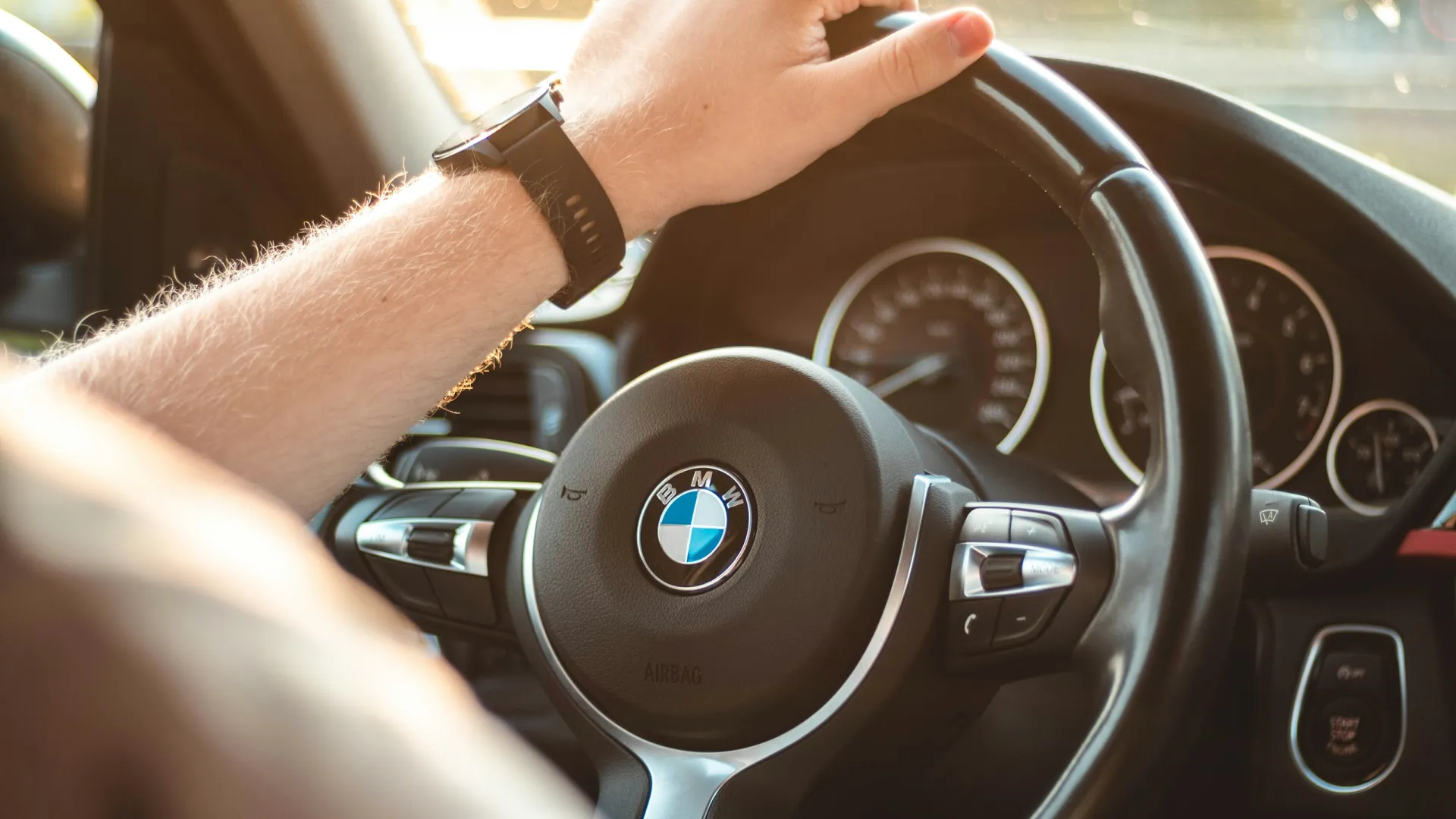 Close-up of a hand on a BMW car steering wheel with dashboard and sunlight in the background