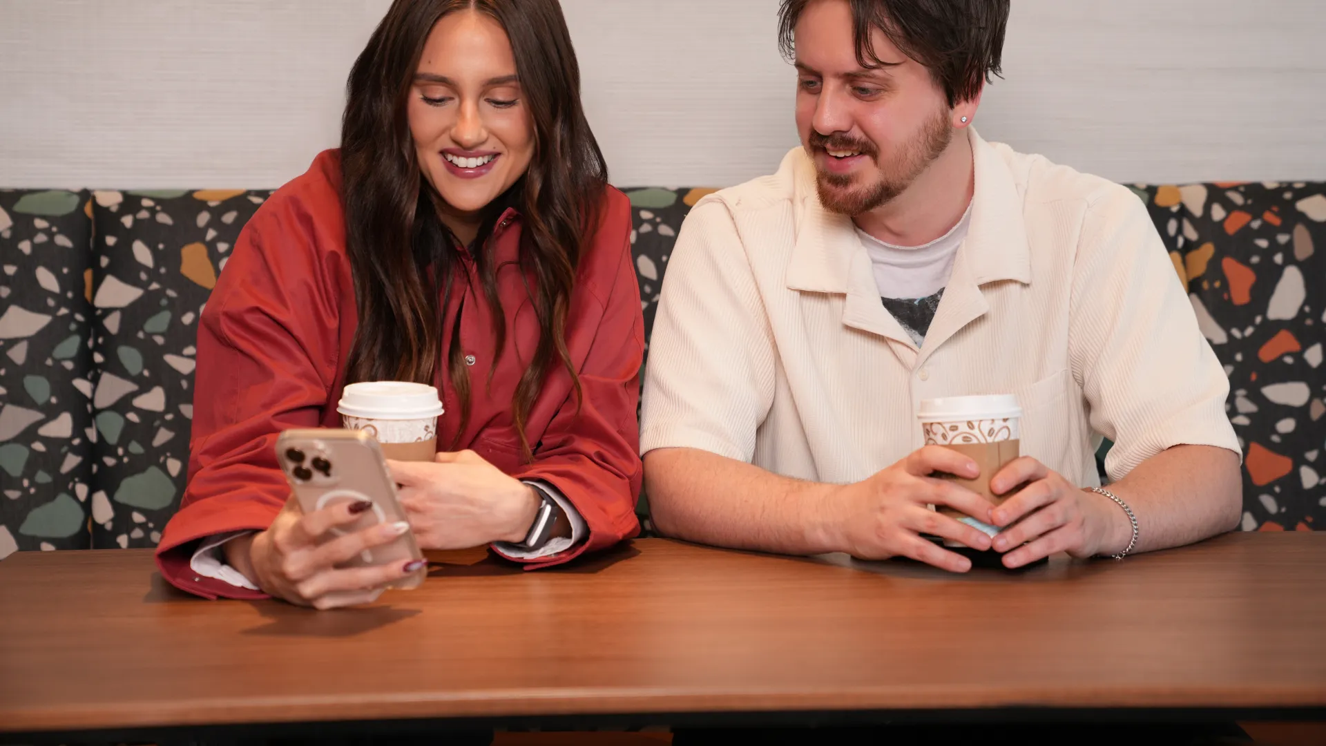 Two young adults smiling and holding coffee cups while looking at a smartphone at a wooden table.