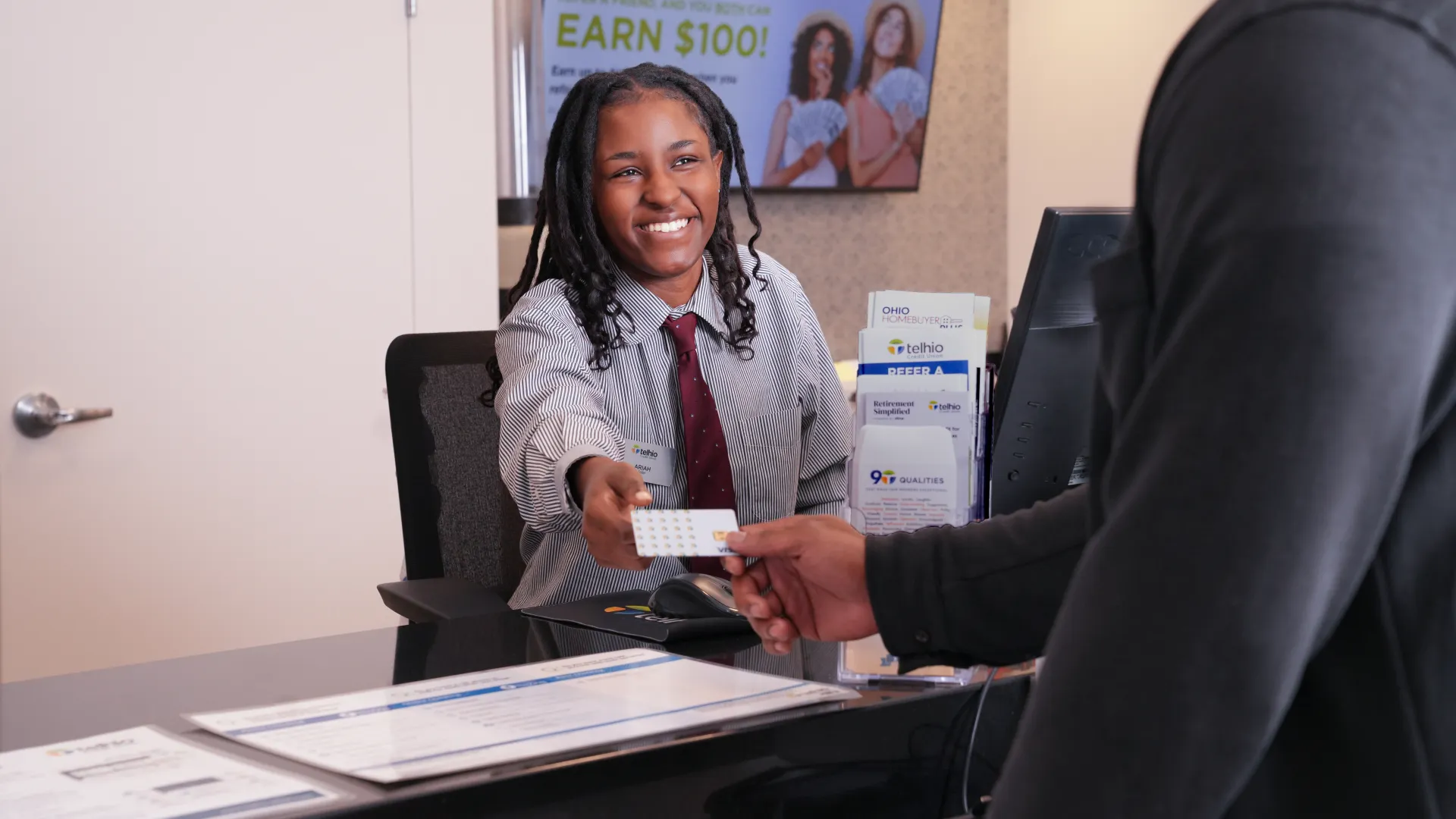 Smiling woman at reception desk hands card to man in dark sweater in bright modern office lobby.