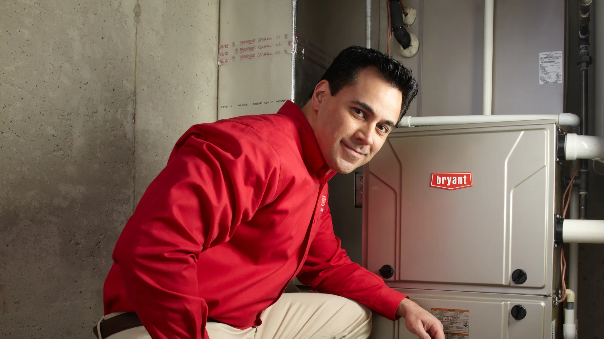 Technician in red shirt kneeling beside a Bryant HVAC unit in a basement setting with tools nearby.