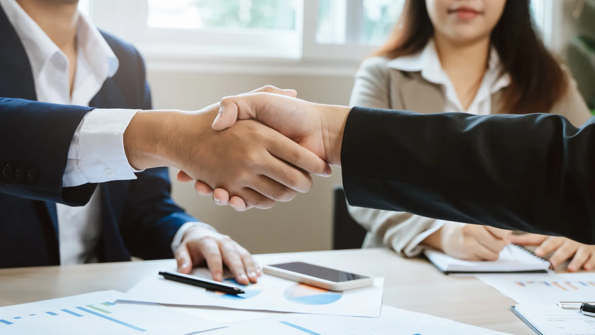 Two business professionals shaking hands over a table with documents and smartphone, signifying agreement.