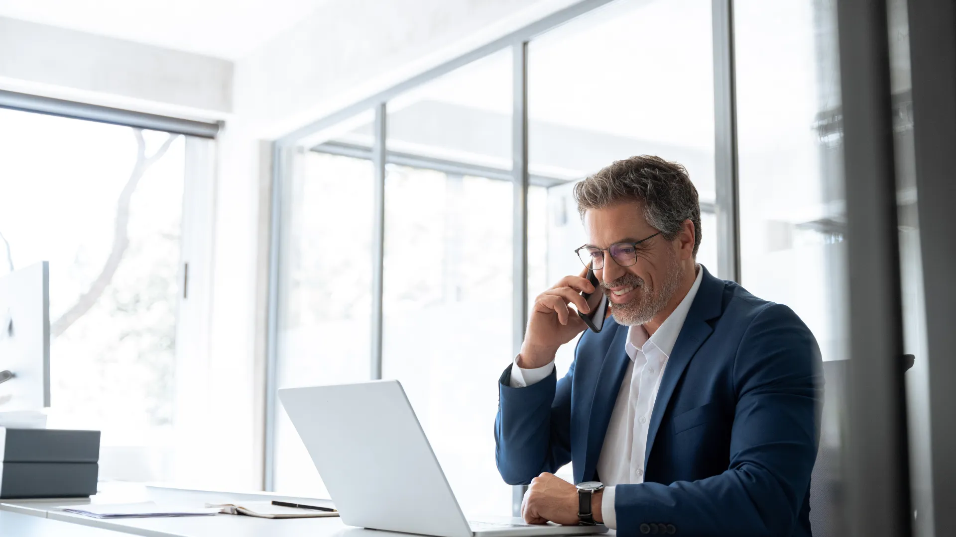 Man in blue suit smiling while talking on phone at desk with laptop in bright modern office