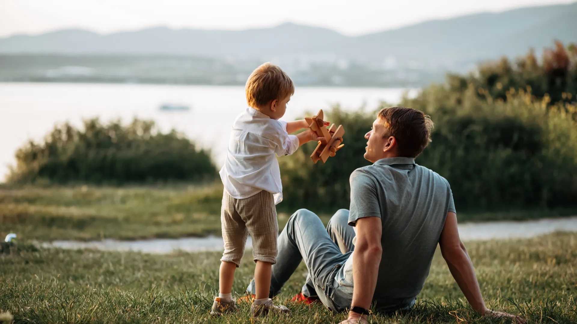 a man and a baby sitting on grass by a body of water