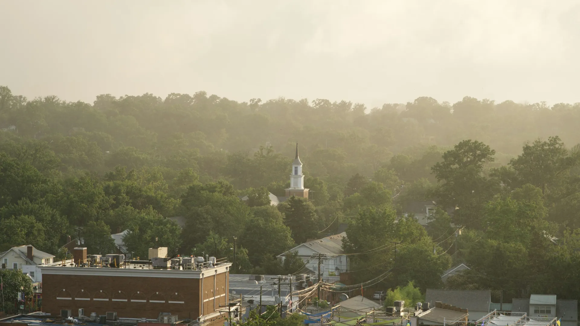 Small town with dense green trees, houses, a church steeple, and hazy sunlight in the background.