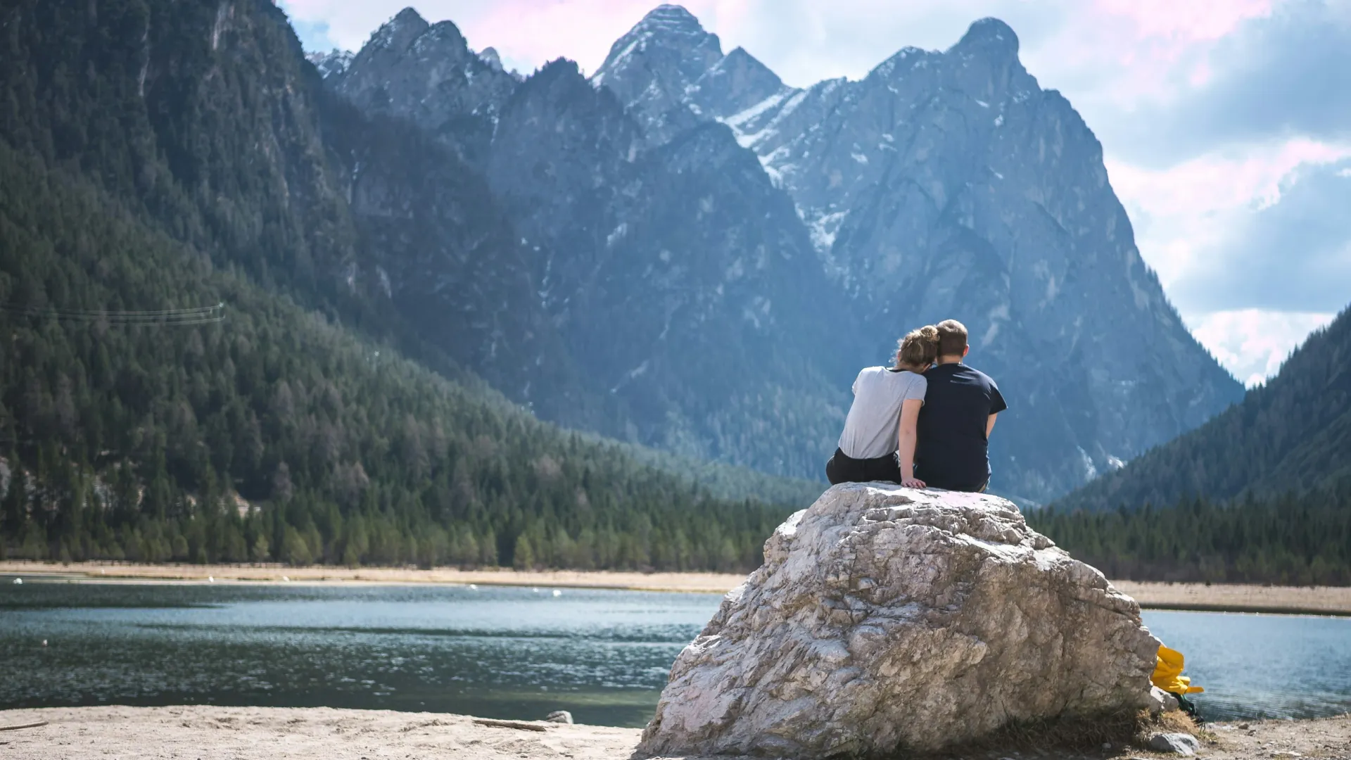 a man and woman sitting on a rock by a lake and mountains