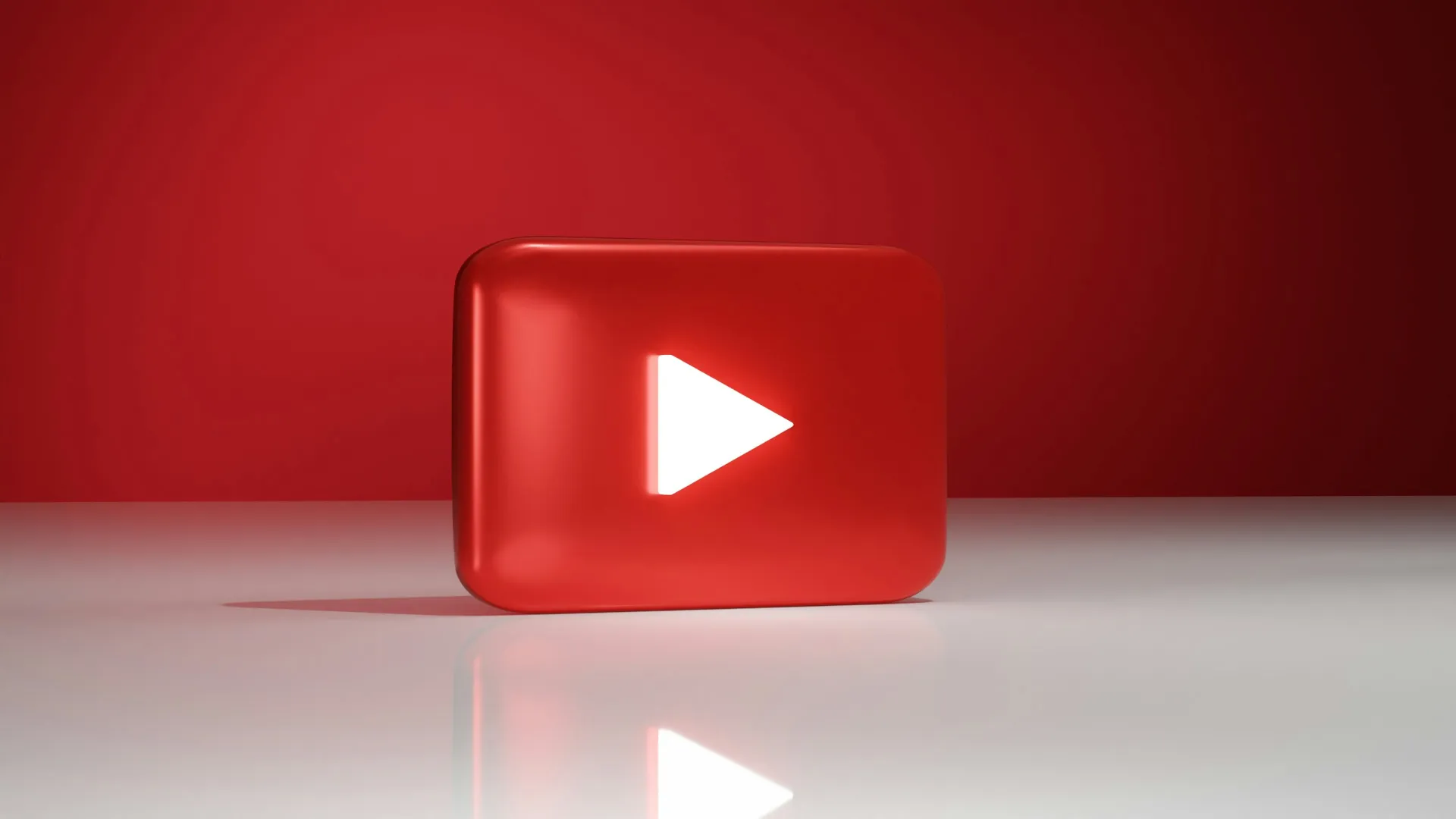 Red 3D YouTube play button logo with white play icon on reflective surface and red background.