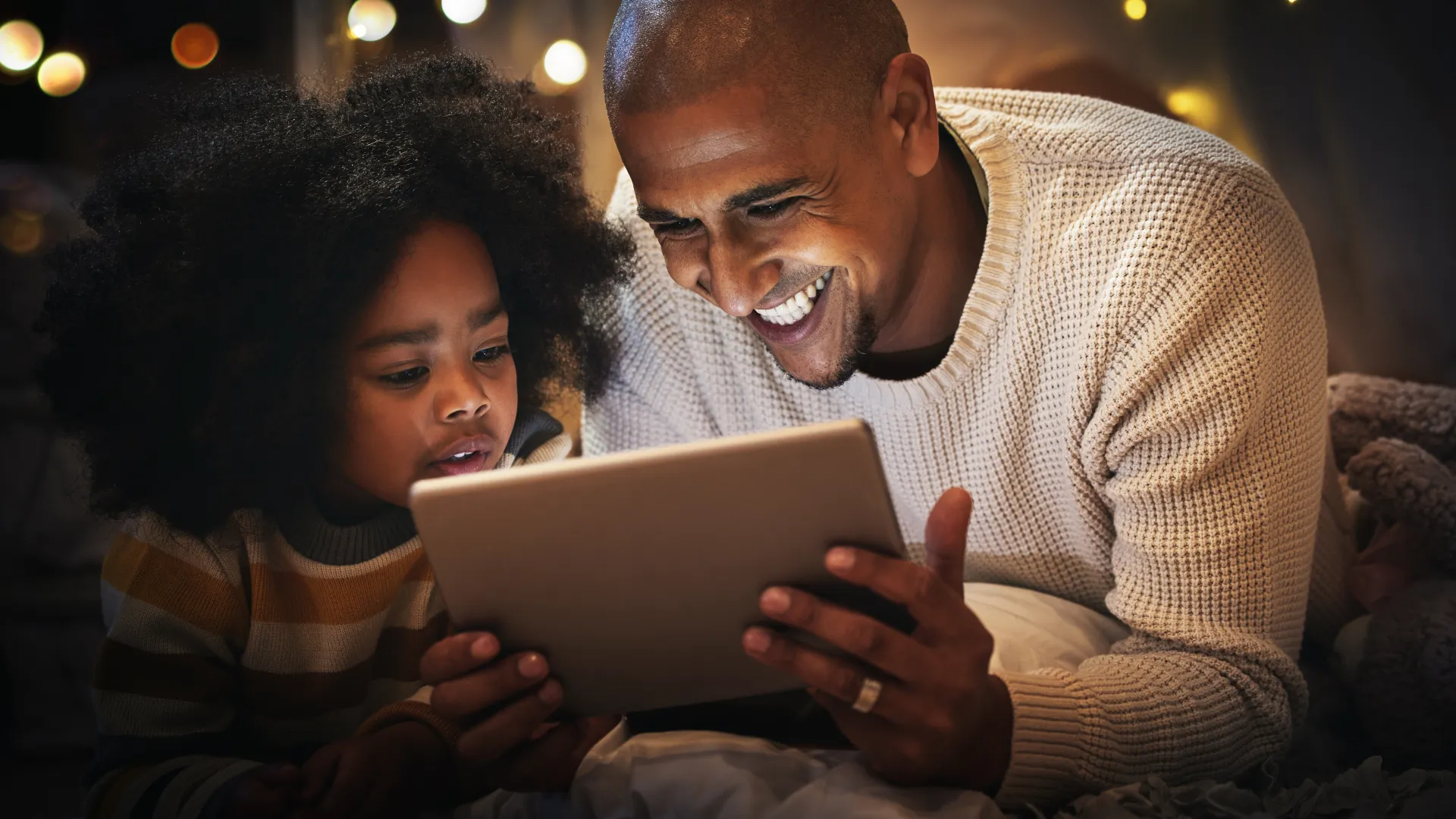 Father and child enjoying a tablet together, surrounded by soft lights and a cozy atmosphere at home.