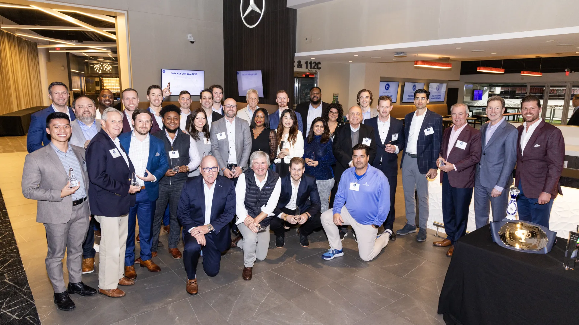 Group photo of diverse professionals posing indoors near large Mercedes-Benz logo at corporate event