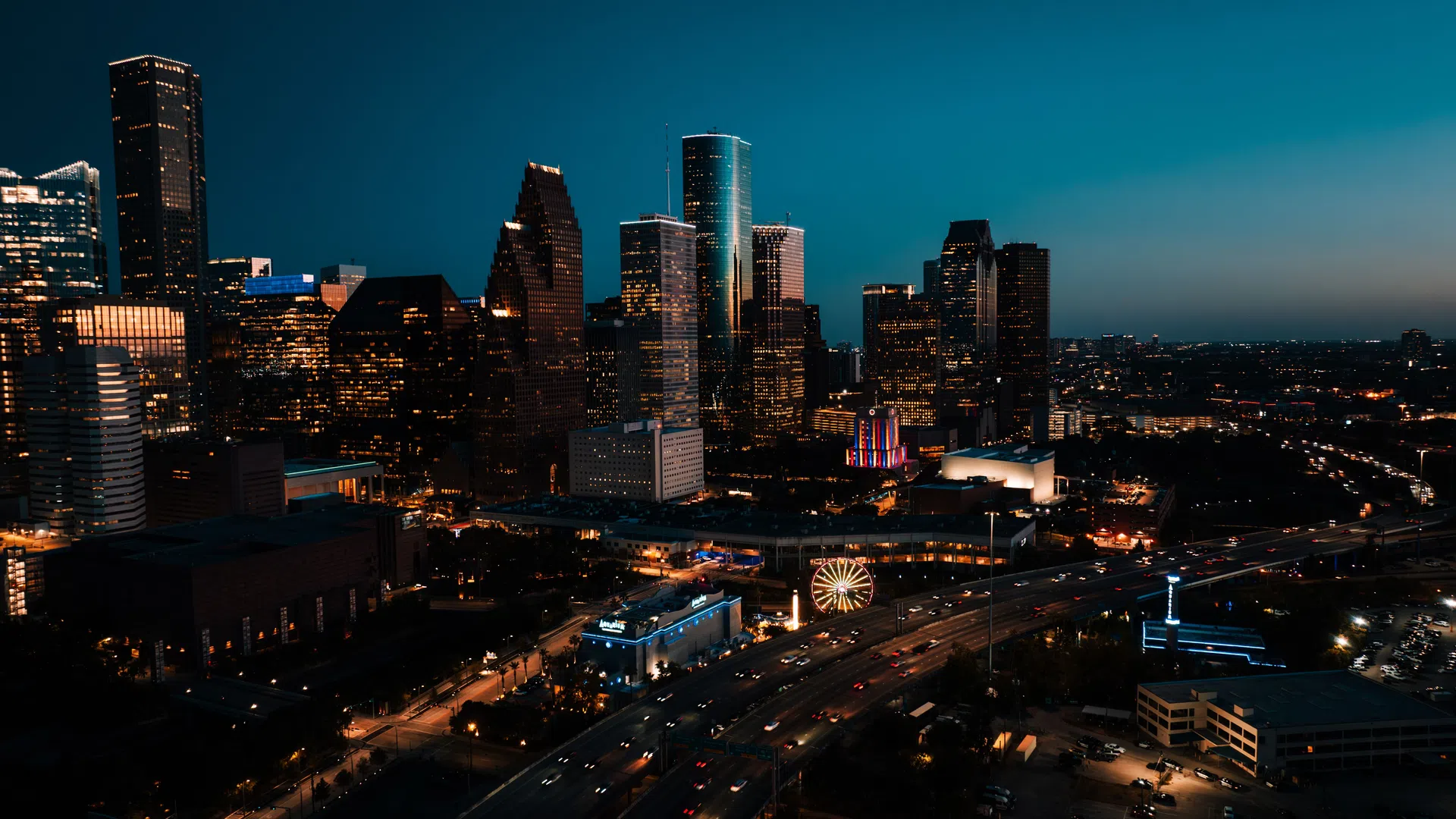 Nighttime cityscape of Houston with illuminated skyscrapers, highways, and a lit Ferris wheel under a dark blue sky.