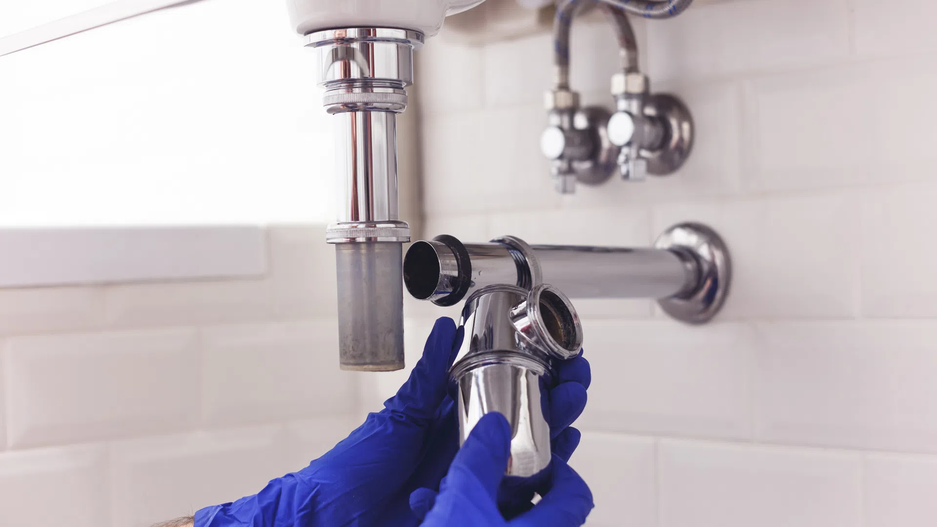 Plumber wearing blue gloves disconnects chrome pipe under a white bathroom sink with tiled wall background
