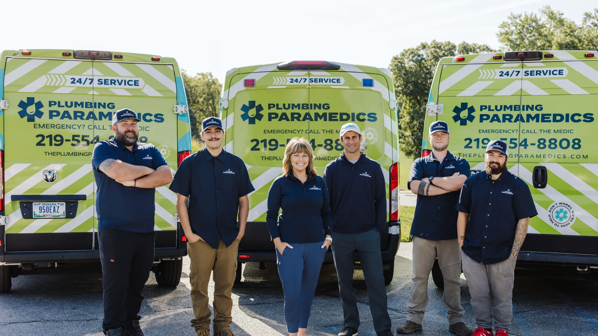 Team of six Plumbing Paramedics technicians standing in front of branded service vans outdoors on sunny day