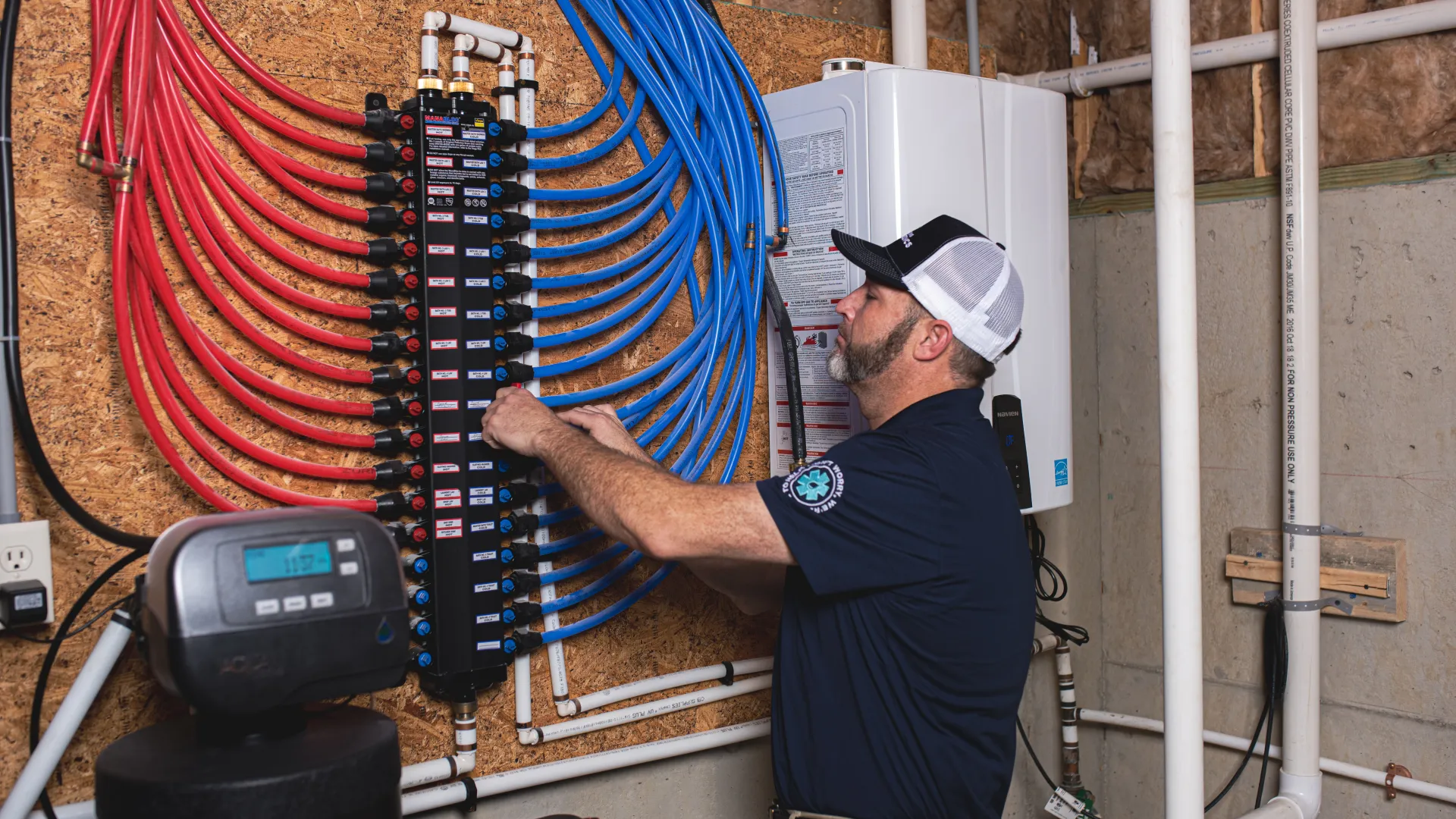 Technician adjusting a manifold with red and blue pipes in a basement utility area beside water heating equipment.