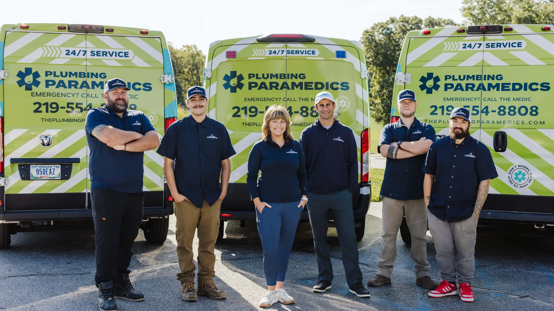 Six Plumbing Paramedics team members stand in front of branded service vans with green and white stripes outdoors.
