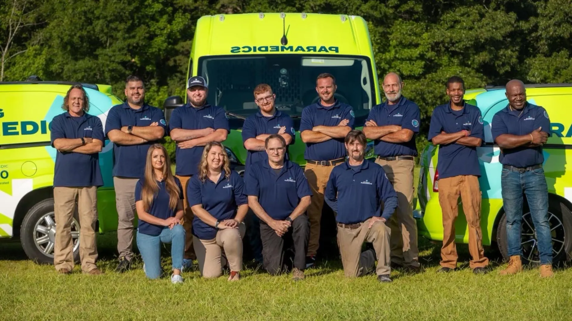 Team of paramedics posing in front of emergency vehicles in a grassy field, showcasing teamwork and professionalism.