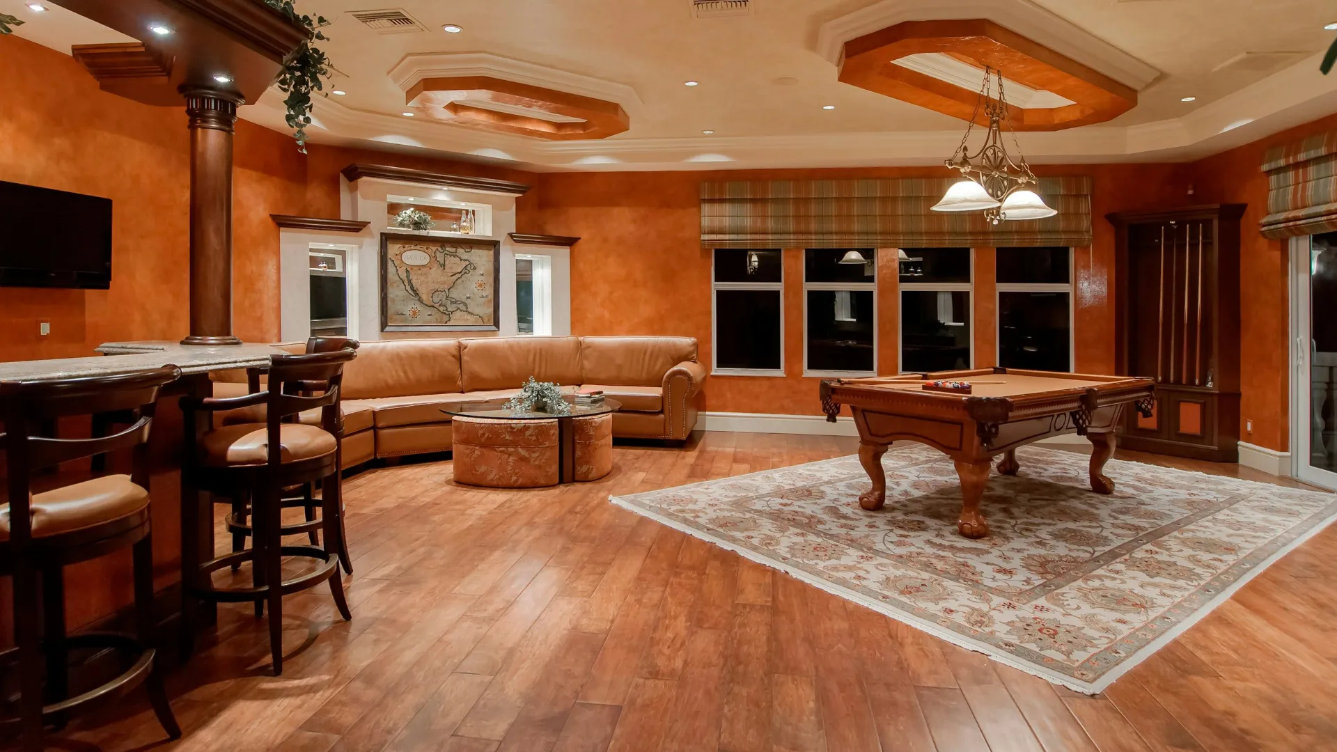 Spacious game room with wooden floors, pool table on patterned rug, leather sectional sofa, and bar seating.
