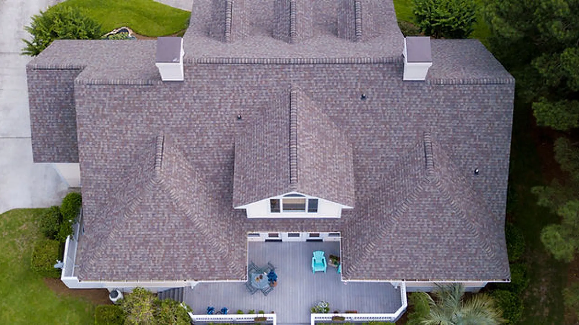 Aerial view of a large house with gray shingled roof surrounded by green trees and lawn on a sunny day