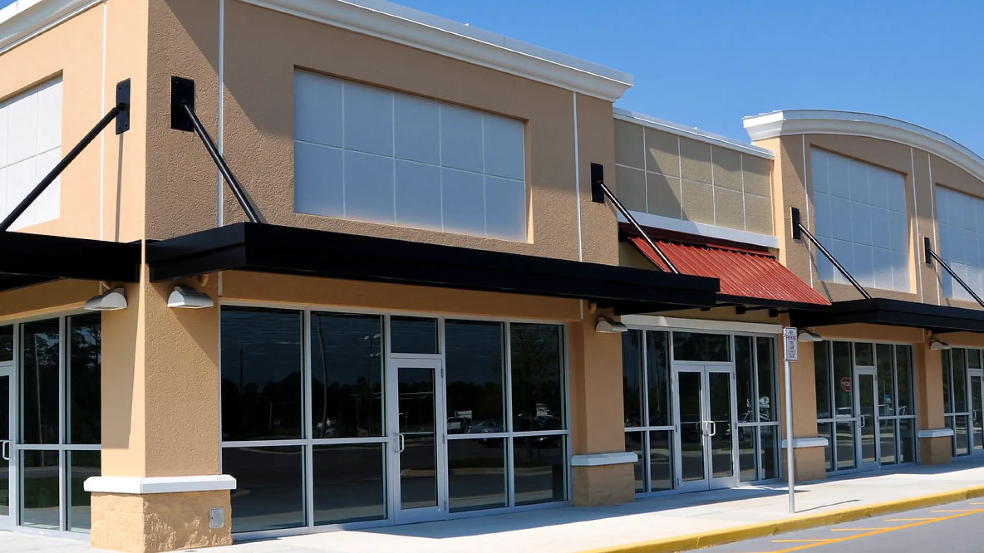 New empty retail shopping plaza with large glass windows and colorful awnings on a sunny day.