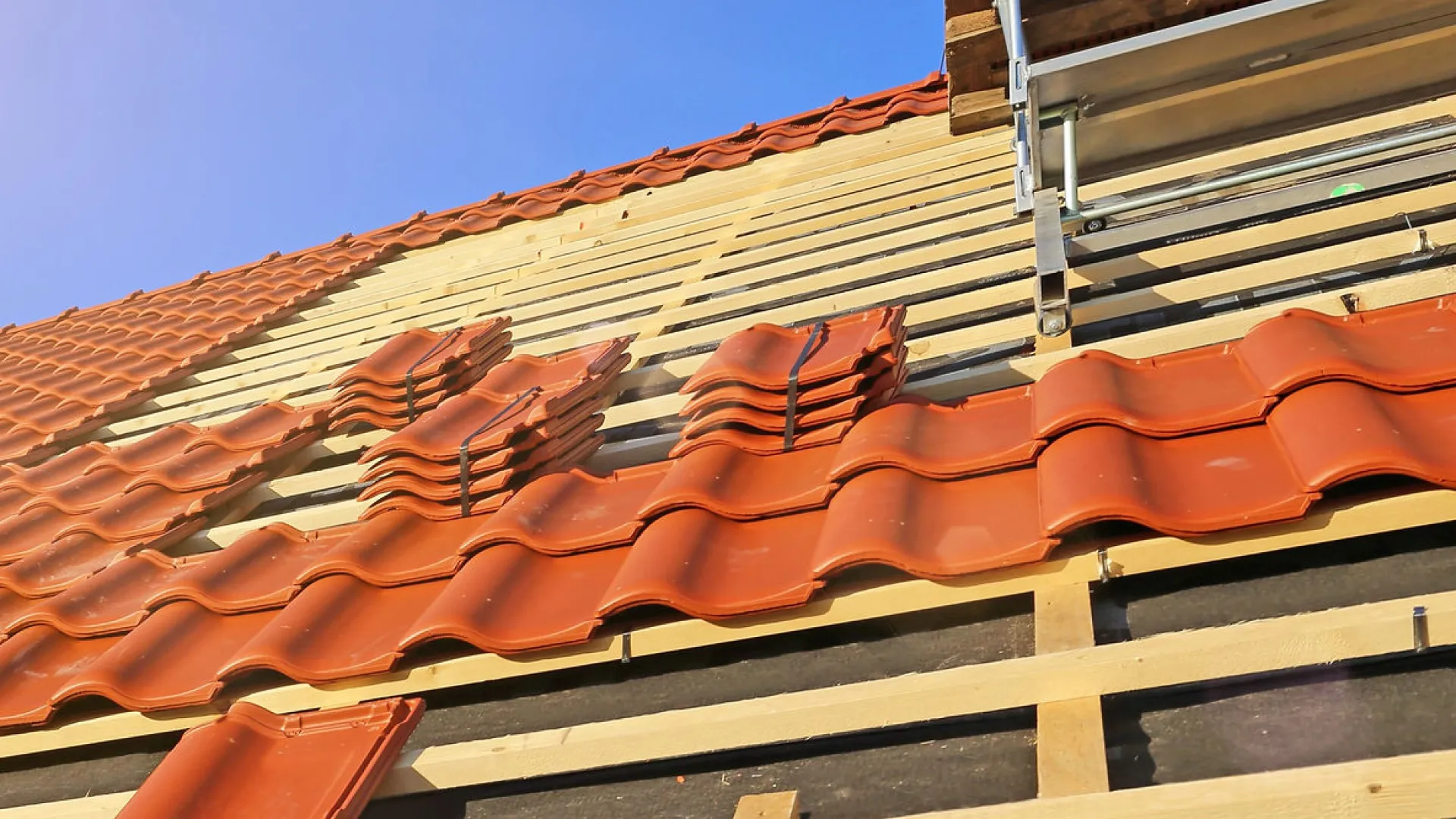 Close-up of terracotta roof tiles being installed on a wooden roof frame under clear blue sky.