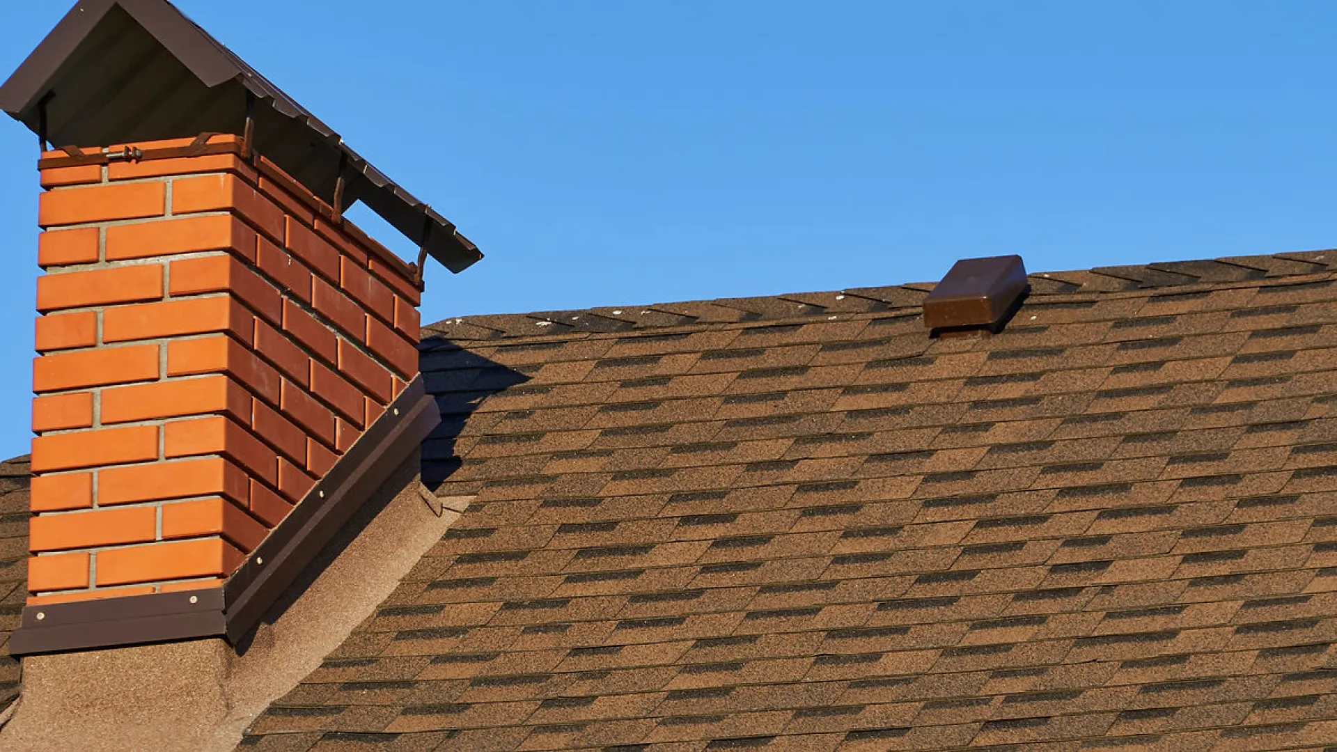 Close-up of brown asphalt shingle roof with brick chimney under clear blue sky.
