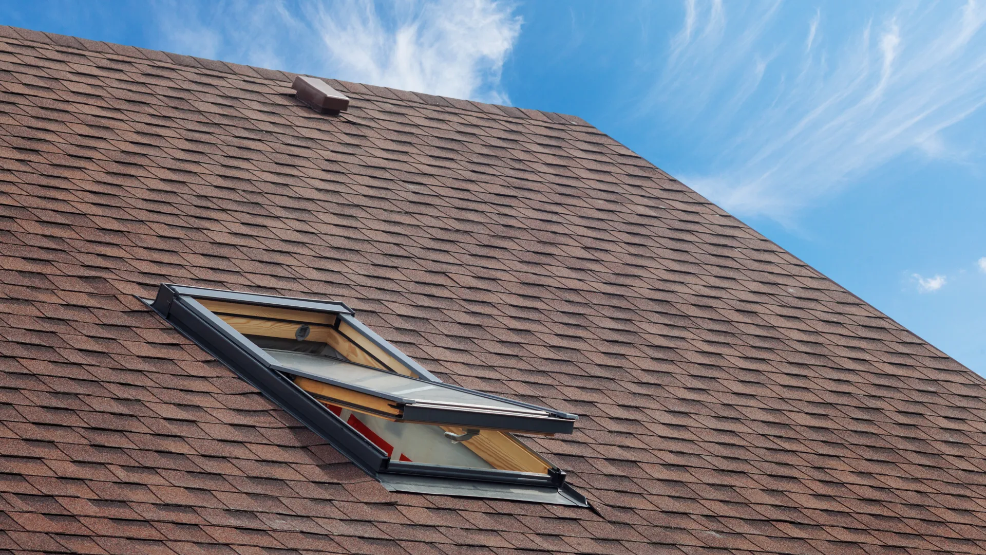 Brown shingled roof with an open skylight window under a blue sky with wispy clouds.