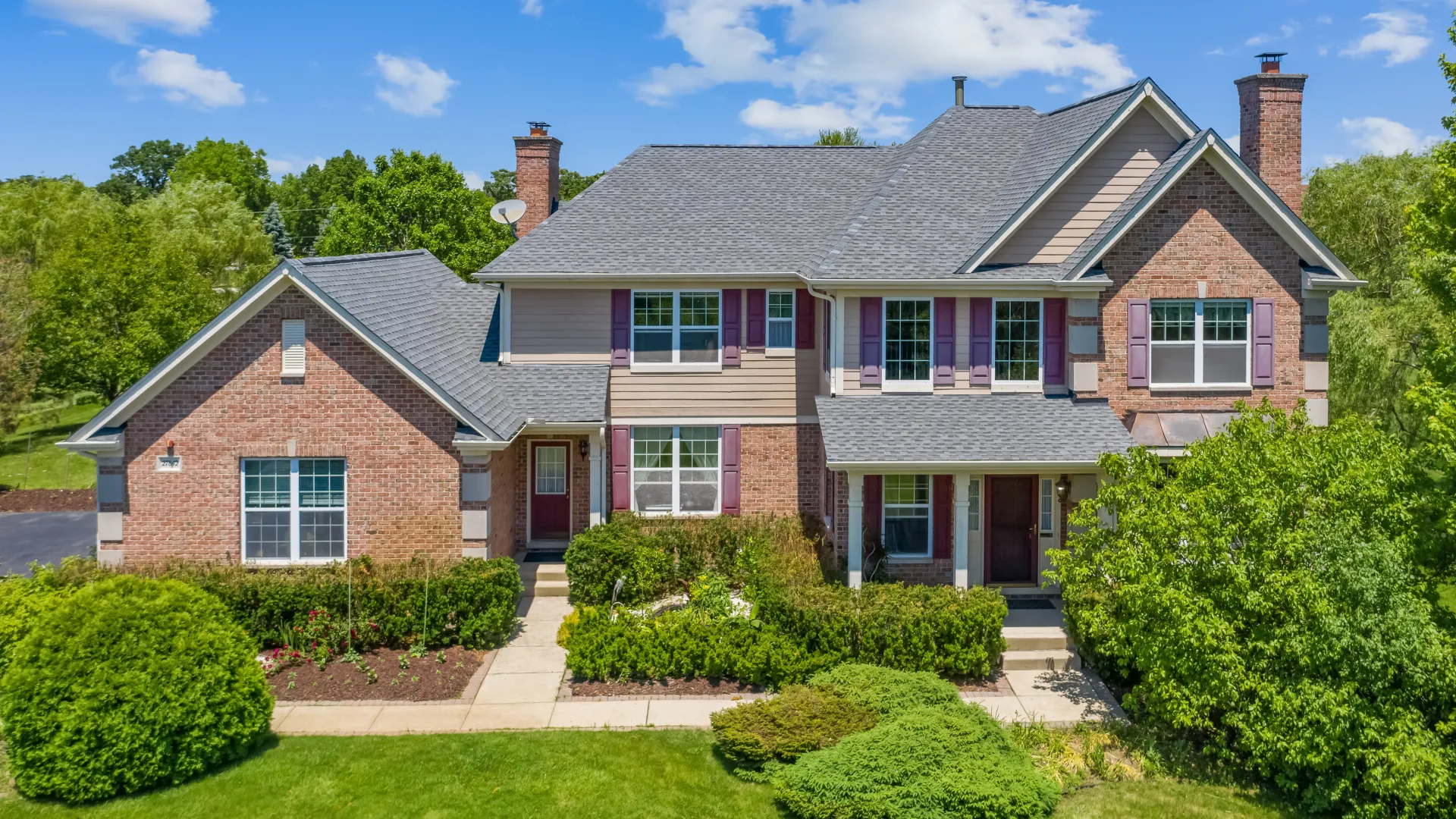 Large two-story suburban house with brick and siding exterior surrounded by lush green landscaping and a blue sky.