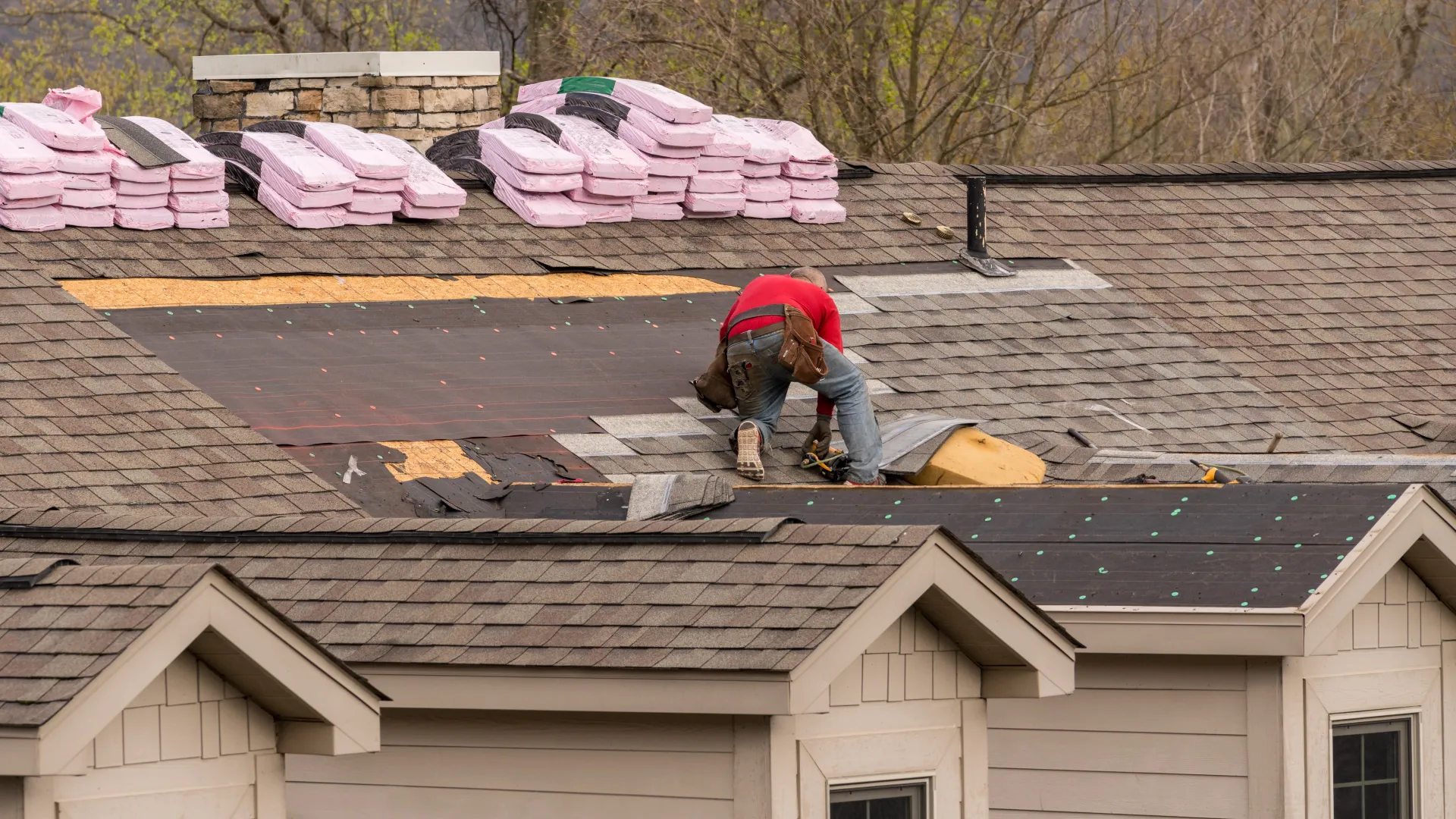 Worker in red shirt installing shingles on a residential roof with roofing materials stacked nearby during daytime.