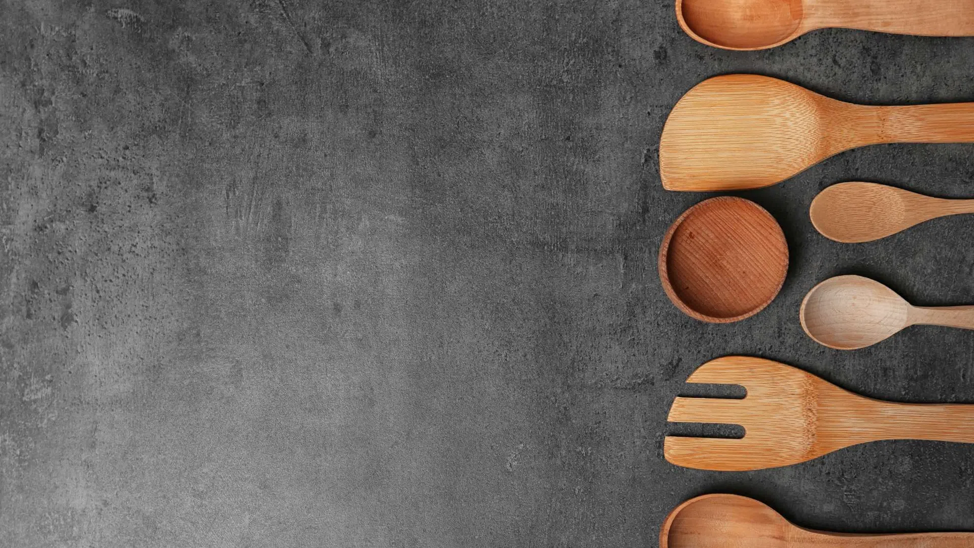 Set of assorted wooden kitchen utensils arranged vertically on a dark textured surface with empty space on the left