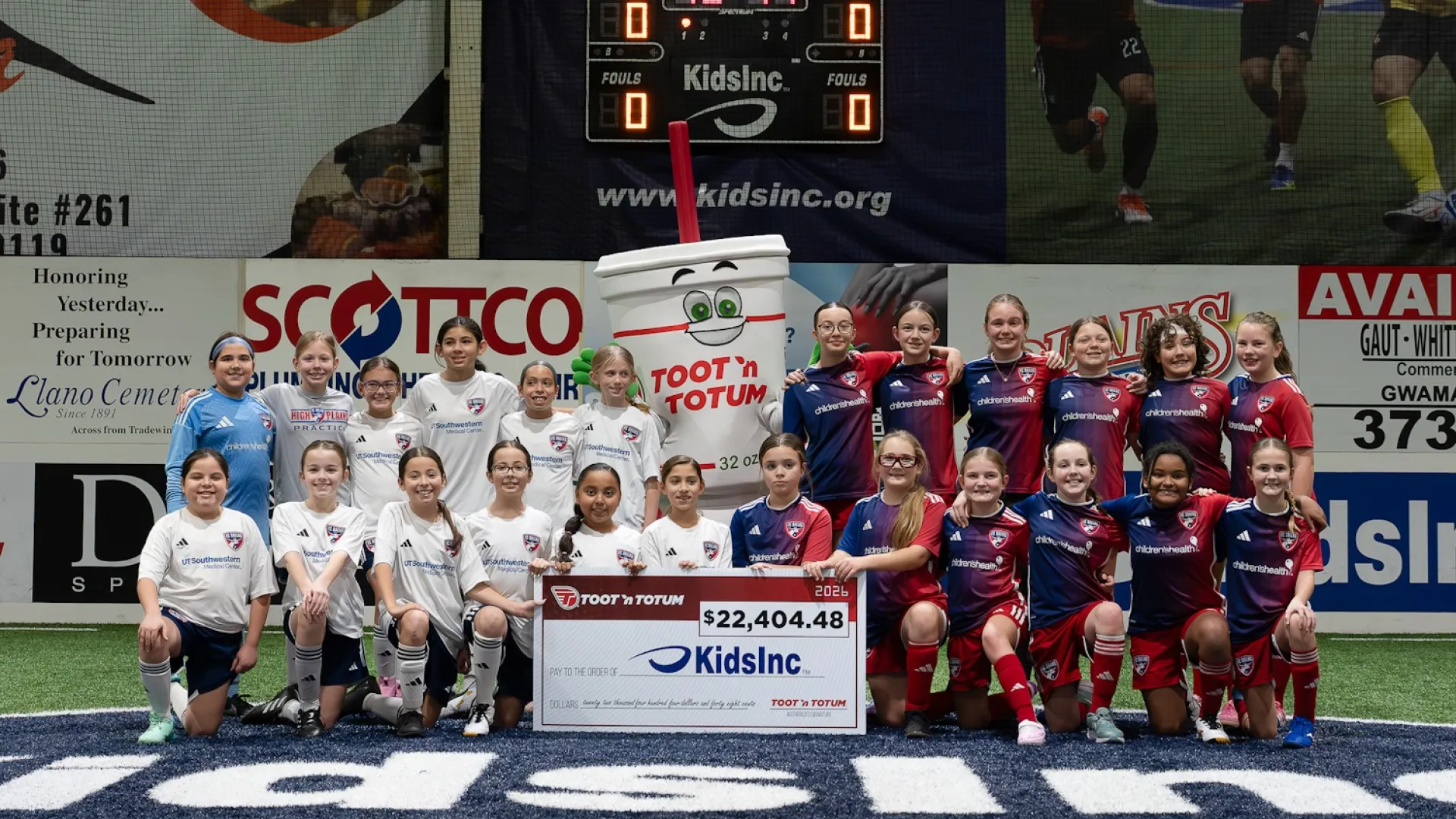 Youth soccer teams pose with mascot and $22,404 donation check for KidsInc on indoor field.