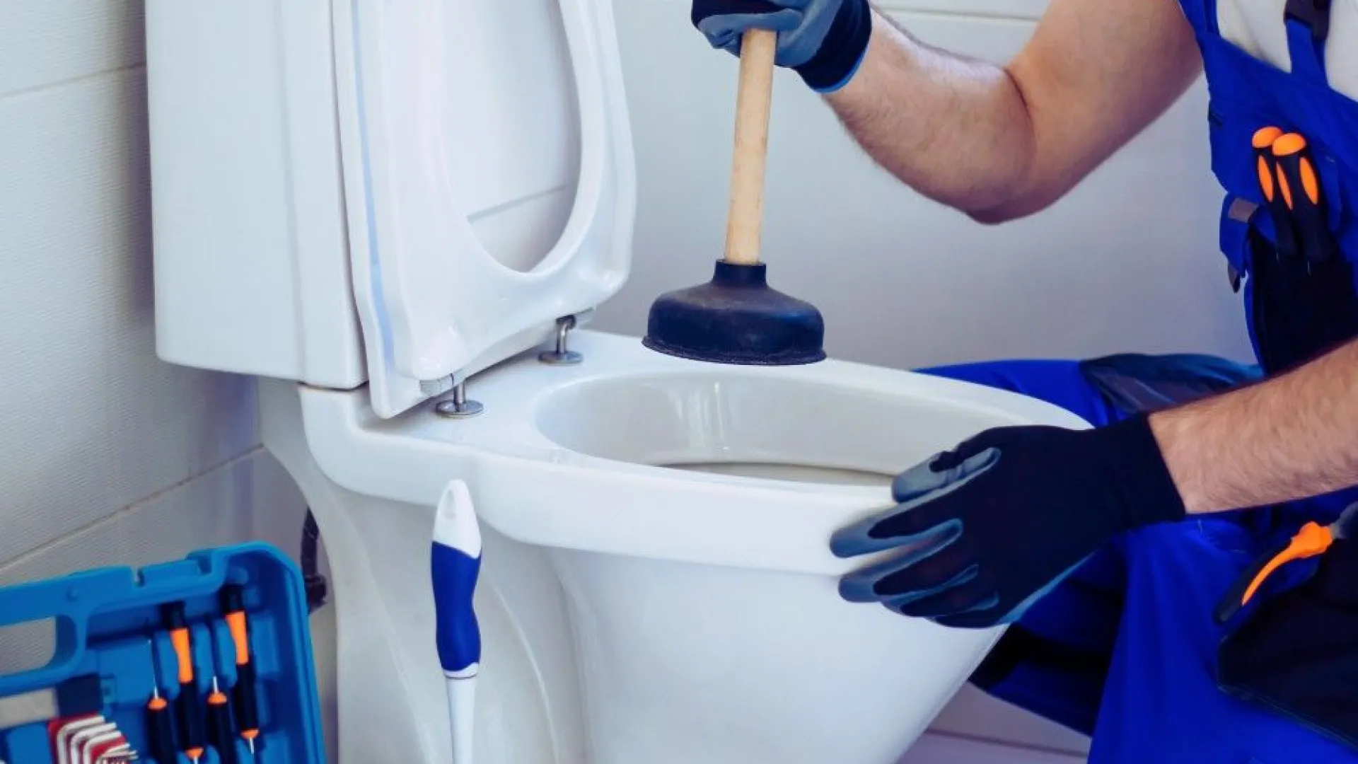 Plumber using a plunger to unclog a white toilet with tools kit nearby in a bathroom.