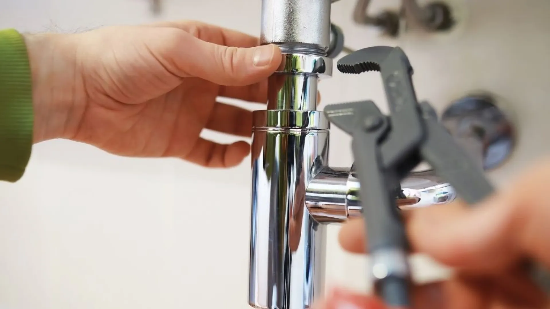 Plumber repairing a shiny chrome pipe under a sink using a wrench with hands in focus.